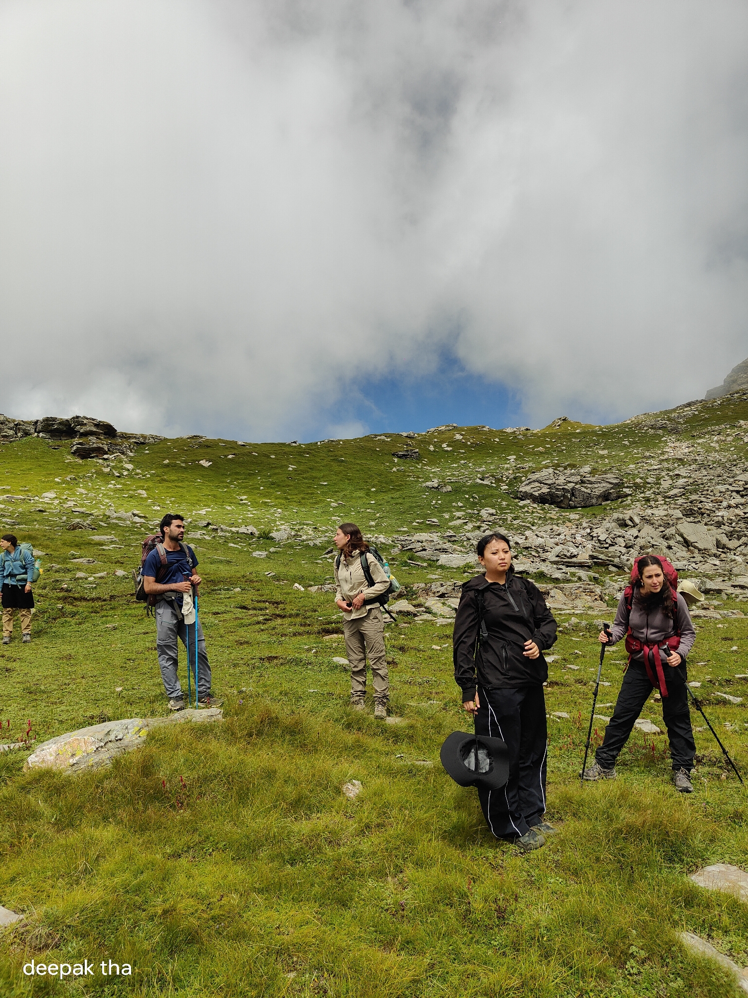The image depicts a group of hikers in a mountainous, grassy terrain, likely in the vicinity of Bashisht, India, given the watermark. The sky is partly cloudy with patches of blue visible. The landscape is characterized by rolling green hills dotted with rocks and boulders.
There are five individuals visible. From left to right:
1. A person in a light blue jacket and dark skirt, facing away from the camera and walking uphill. They have a backpack.
2. A man in a blue t-shirt and grey pants, with a backpack, holding two trekking poles. He is looking to his right and appears to be in mid-conversation.
3. A person in a light brown jacket and beige pants, with a backpack, facing the camera and looking slightly to their left.
4. A person in a black jacket and black pants with white stripes, carrying a black hat in their left hand and holding a trekking pole in their right. They are looking forward and slightly to their right.
5. A person in a purple jacket and black pants, with a red backpack, holding two trekking poles and looking forward and slightly to their left.
The individuals are equipped for hiking, with backpacks and trekking poles. The scene conveys a sense of outdoor activity and exploration in a natural, rugged environment.