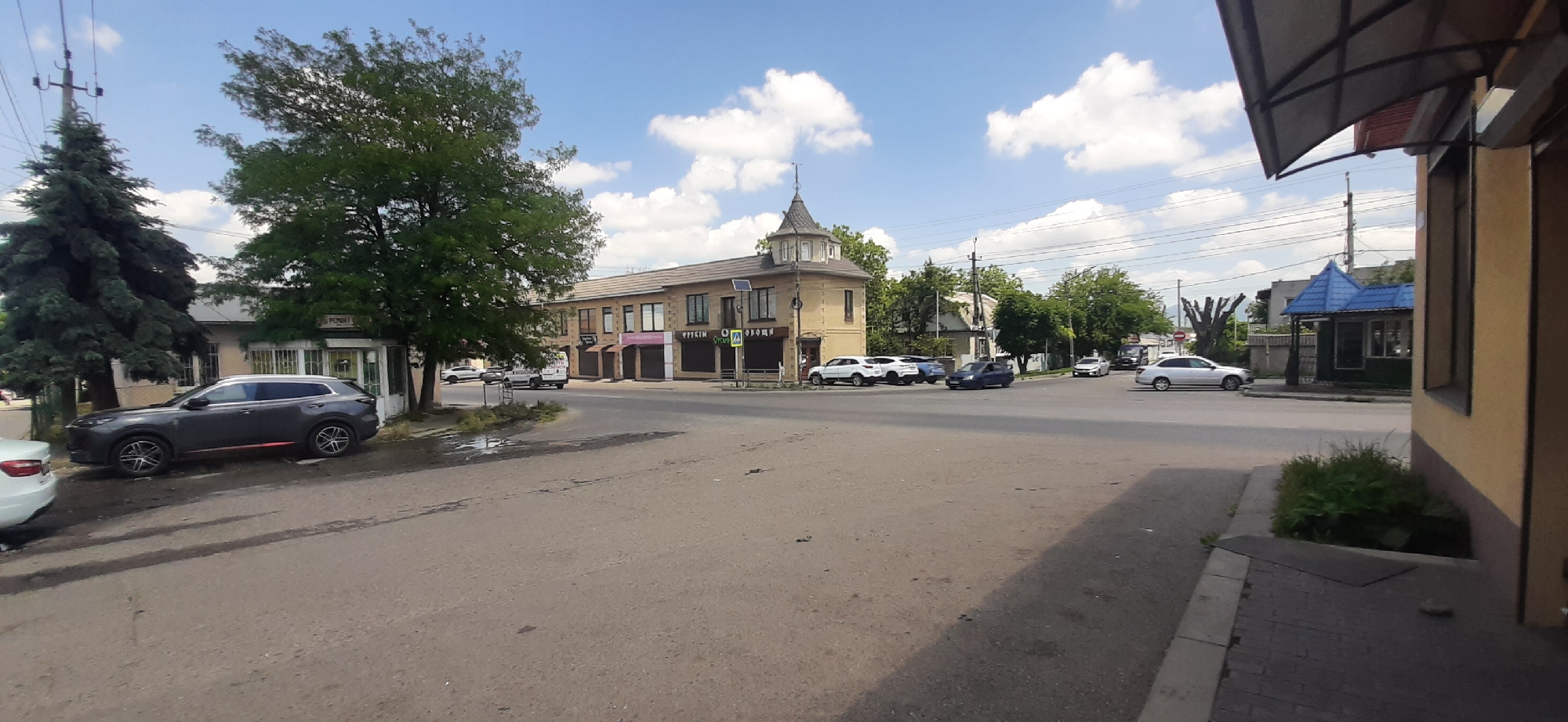 The image depicts a street scene in Yessentukskaya, Russia, on a sunny day with scattered clouds. In the foreground, a paved road is visible, with a sidewalk on the right side. A dark grey SUV is parked on the left side of the road. Behind the SUV, a building with a light-colored facade and windows is partially visible, with a green Christmas tree and another deciduous tree in front of it.
Further down the street, a light-yellow building with multiple windows and shops on the ground floor is prominent. This building has a distinctive tower-like structure with a pointed roof. Several cars are parked in front of and near this building, including a white SUV, a dark blue sedan, and other vehicles. A traffic sign with a pedestrian symbol is visible on a pole in front of the building.
On the right side of the image, a smaller building with a blue, peaked roof is situated near the street. Further back, trees and other buildings can be seen. The overall impression is that of a moderately busy street in a town or small city, with commercial establishments and residential areas mixed.