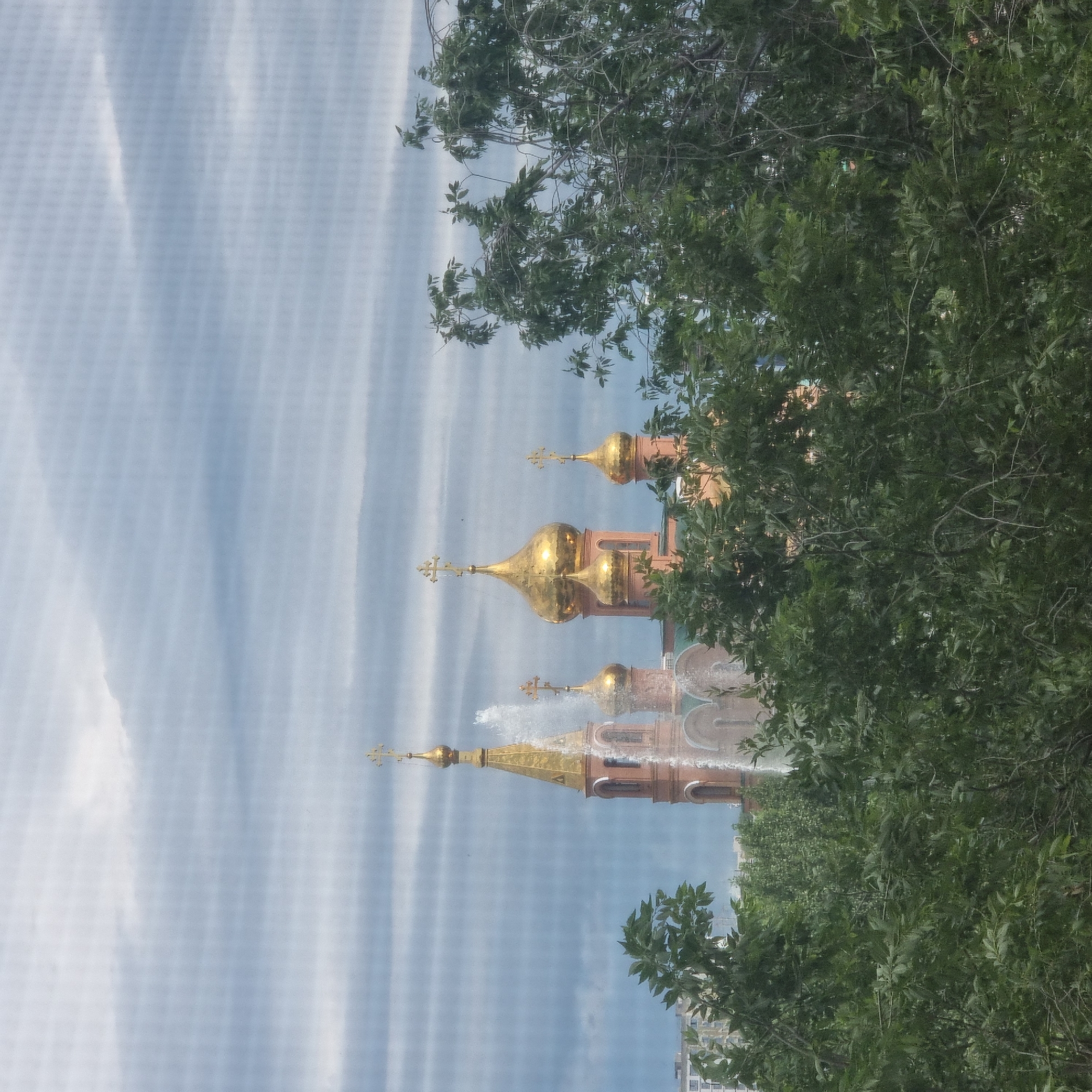 The image depicts an Orthodox church with golden domes and crosses, partially obscured by lush green trees in the foreground. A fountain is visible to the left of the church, with water spraying upwards. The sky is overcast with soft, horizontal cloud formations. The church appears to be situated in an urban environment, with some buildings visible in the background to the far left. This scene is consistent with the location of the Church of the Assumption of the Virgin Mary in Aktobe, Kazakhstan, which features similar architectural elements and is surrounded by greenery. There are no people or animals visible in the frame.