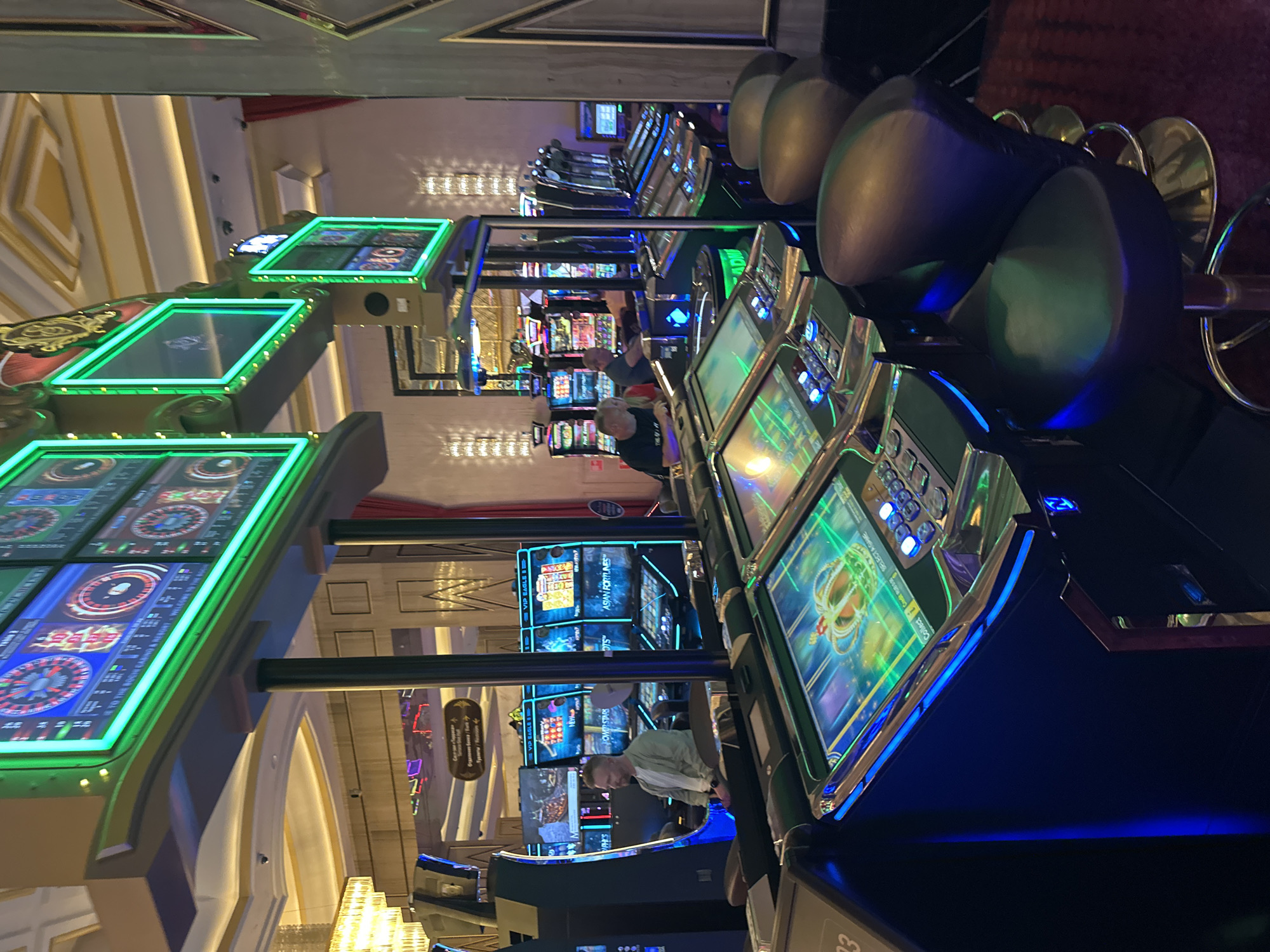 The image shows a brightly lit casino floor with multiple slot machines. In the foreground, a row of slot machines with illuminated screens display colorful graphics, including a golden crown. The machines have a sleek, modern design with blue and green neon accents. Several gaming stools with black leather seats and polished chrome bases are positioned in front of the machines.
In the mid-ground, a man with short blonde hair and a light-colored shirt is seated at a slot machine, appearing to be playing. Further back, other slot machines are visible, some with people seated at them. The casino environment is characterized by ornate ceilings with gold detailing and chandeliers, suggesting a luxurious setting. The overall impression is of a bustling casino in an indoor environment. The text on one of the screens indicates "VIP EAGLE" and "ASIAN FORTUNES", suggesting specific game titles. A directional sign with Cyrillic text is also visible in the background.
