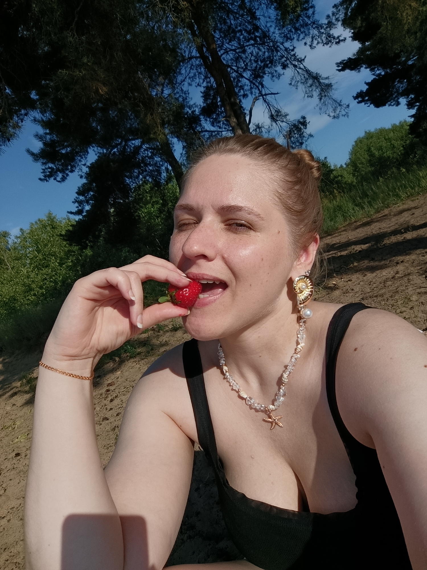 A young woman with her hair in a bun is taking a selfie outdoors. She is wearing a black tank top and jewelry, including a bracelet, necklace with a starfish pendant, and an earring with a pearl. She is holding a strawberry to her mouth, with her eyes closed and a slight smile. The background is a sandy area with trees and a blue sky, suggesting a natural or park-like setting. The lighting indicates it is daytime.