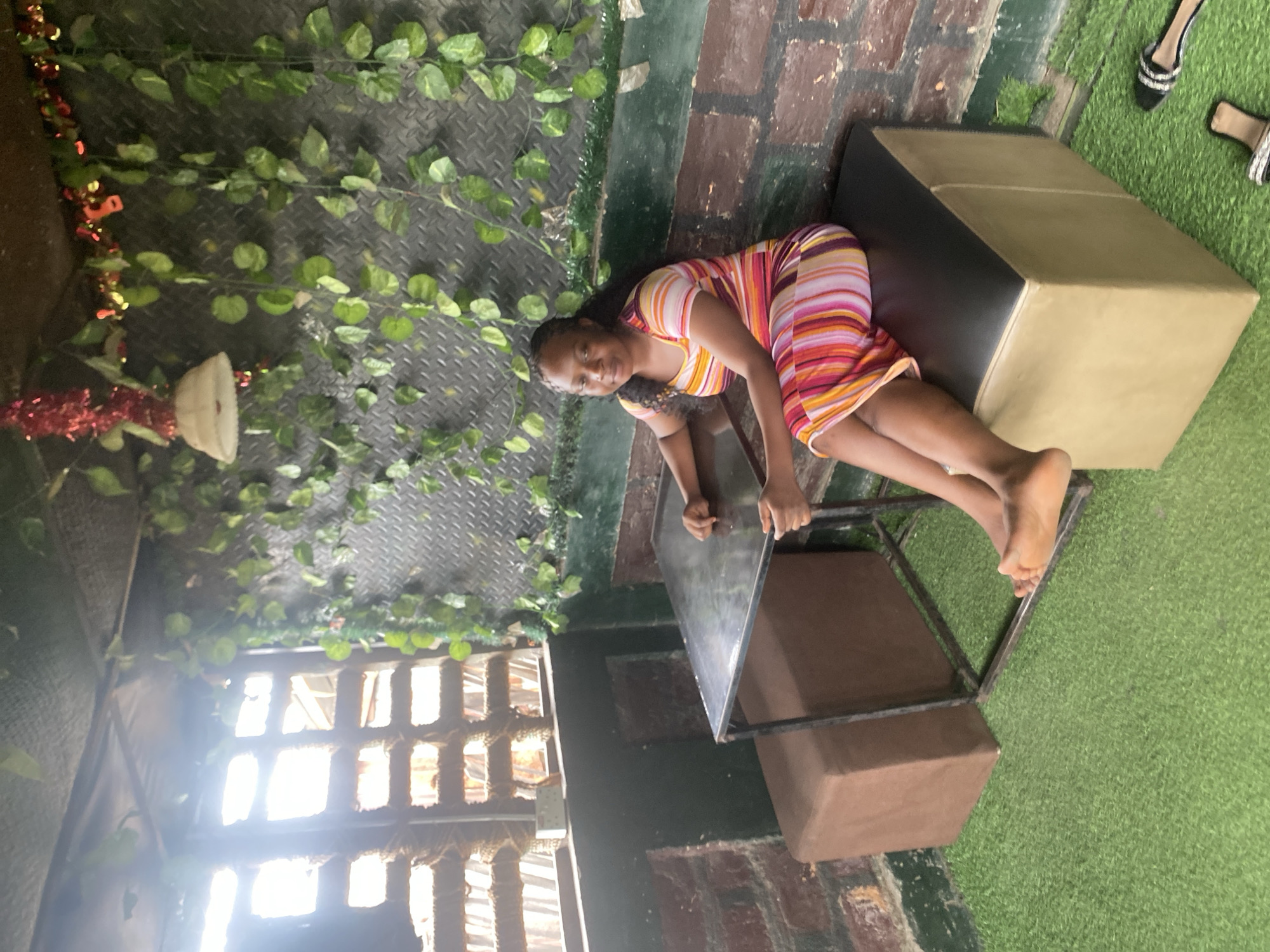A young Black woman with braided hair is posing on a multi-level metal and brown upholstered structure in an outdoor setting. She is wearing a brightly striped dress with pink, yellow, white, and purple stripes. The woman is lying on her side, looking towards the camera with a slight smile. The structure appears to be made of metal frames and upholstered boxes, with one box being a dark gray and the other a tan color. The ground is covered in artificial green turf, and there are some decorative vines with green leaves attached to a diamond-patterned metal wall in the background. Red and gold tinsel-like decorations are also visible among the vines. To the right of the woman, a pair of black heels with crystal embellishments and a tan sandal are visible on the turf. In the background, there is a window with a wooden lattice, through which sunlight streams in, suggesting it is daytime. The overall scene has an eclectic, possibly playful, or decorative feel. No text is visible in the image.