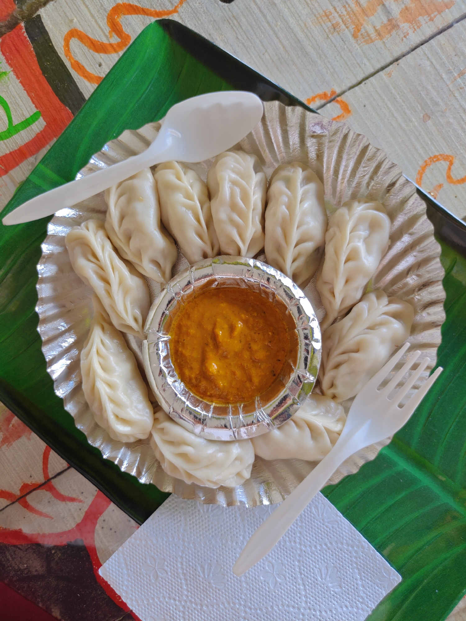 A close-up, overhead shot showcases a serving of momos, a popular Nepali dumpling, arranged on a disposable aluminum foil plate. The momos, a pale cream color and neatly folded, encircle a small, circular container filled with a thick, orange-brown dipping sauce. A white plastic spoon rests on the left side of the plate, and a white plastic fork is positioned on the right. The plate is placed on a green banana leaf, which in turn is on a table with a patterned surface featuring red, orange, and black abstract designs. A white paper napkin with a floral embossed pattern is situated in the lower right corner, partially beneath the fork. The scene appears to be set in an outdoor or casual dining environment, likely a street food stall or a simple eatery within Kathmandu Metropolitan City, Nepal, given the cultural context of momos. There are no people visible in the frame.