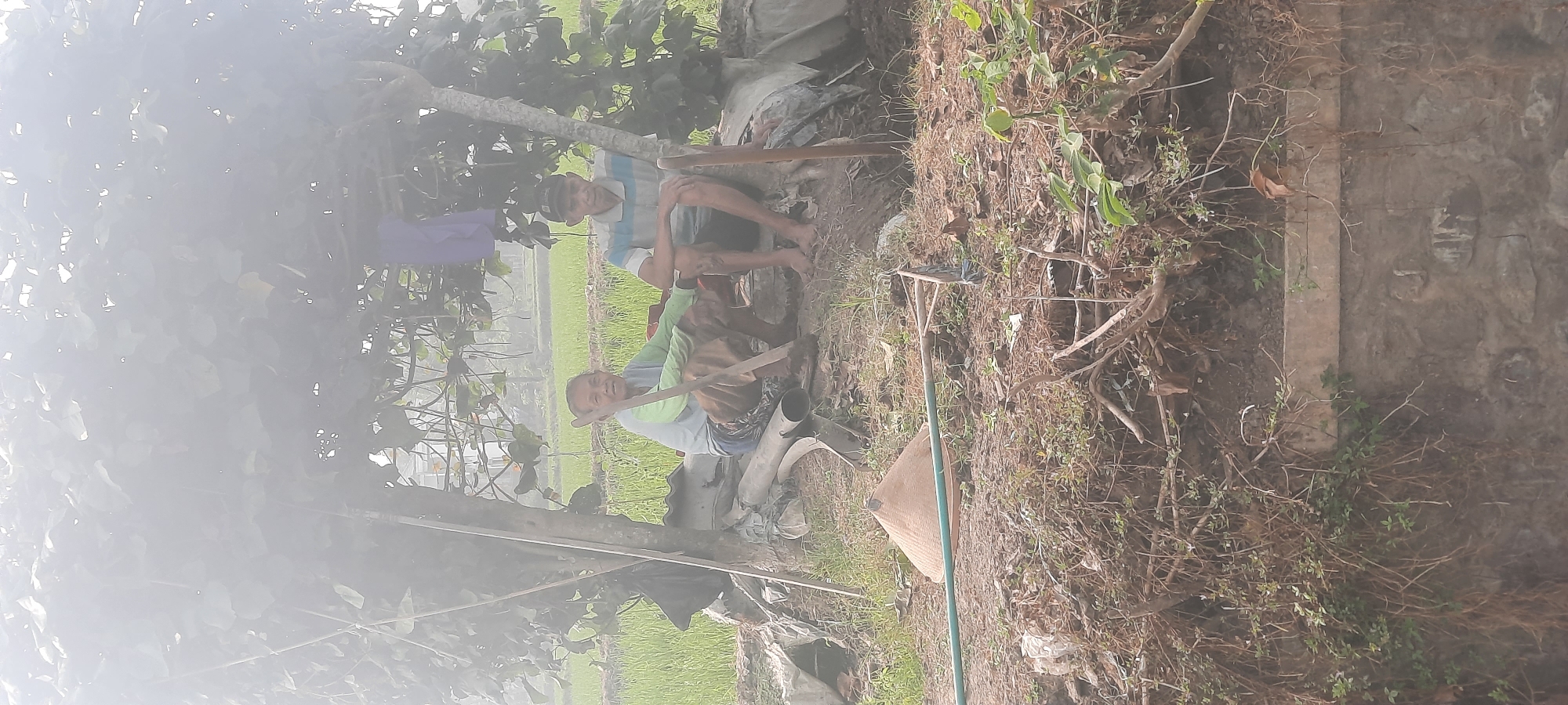 Two individuals are seated under a canopy of leaves, in what appears to be a rural setting near Ploso, Indonesia. The person on the left, an older woman wearing a light blue long-sleeved shirt and patterned pants, is holding a wooden tool. She is looking towards the camera. To her right, a man wearing a striped short-sleeved shirt and a dark baseball cap is also seated, facing forward. They are positioned on a dirt surface with various items around them, including a conical hat, pipes, and other discarded materials. In the background, a lush green rice field stretches out, indicating an agricultural area. The scene is somewhat obscured by a hazy, diffused light, possibly indicating early morning or a misty day.