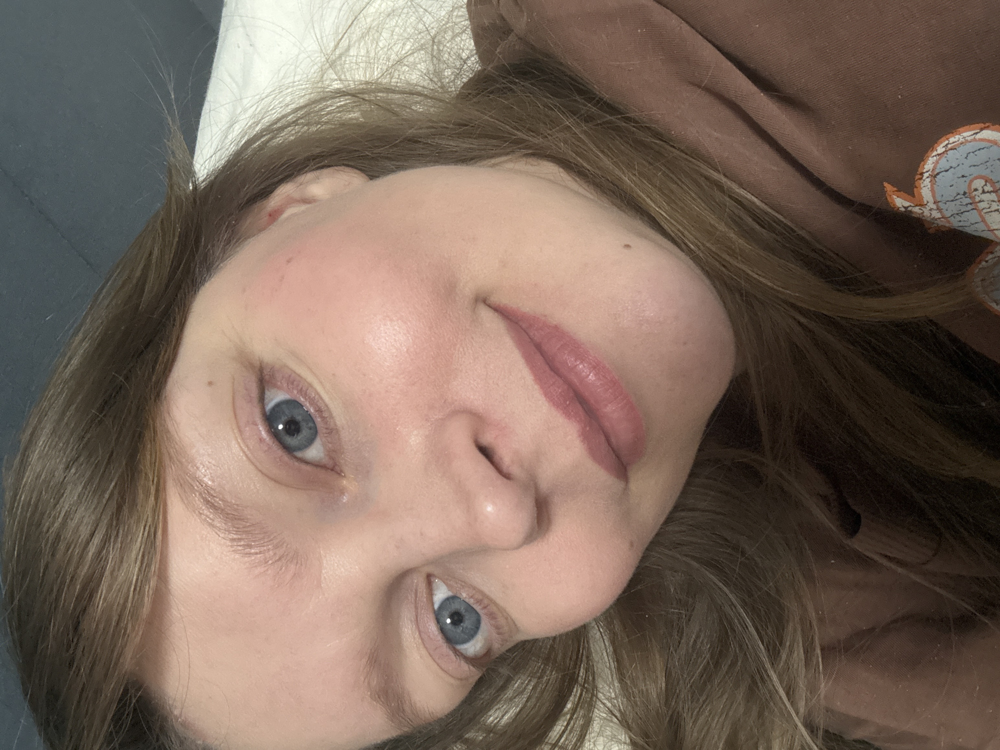 The image shows a close-up selfie of a fair-skinned woman with light brown hair and blue eyes. She is wearing a brown hoodie. Her head is tilted to the left. The background includes a white pillow and a blue wall or surface. There is no information to confirm the location is Izhevsk, Russia.