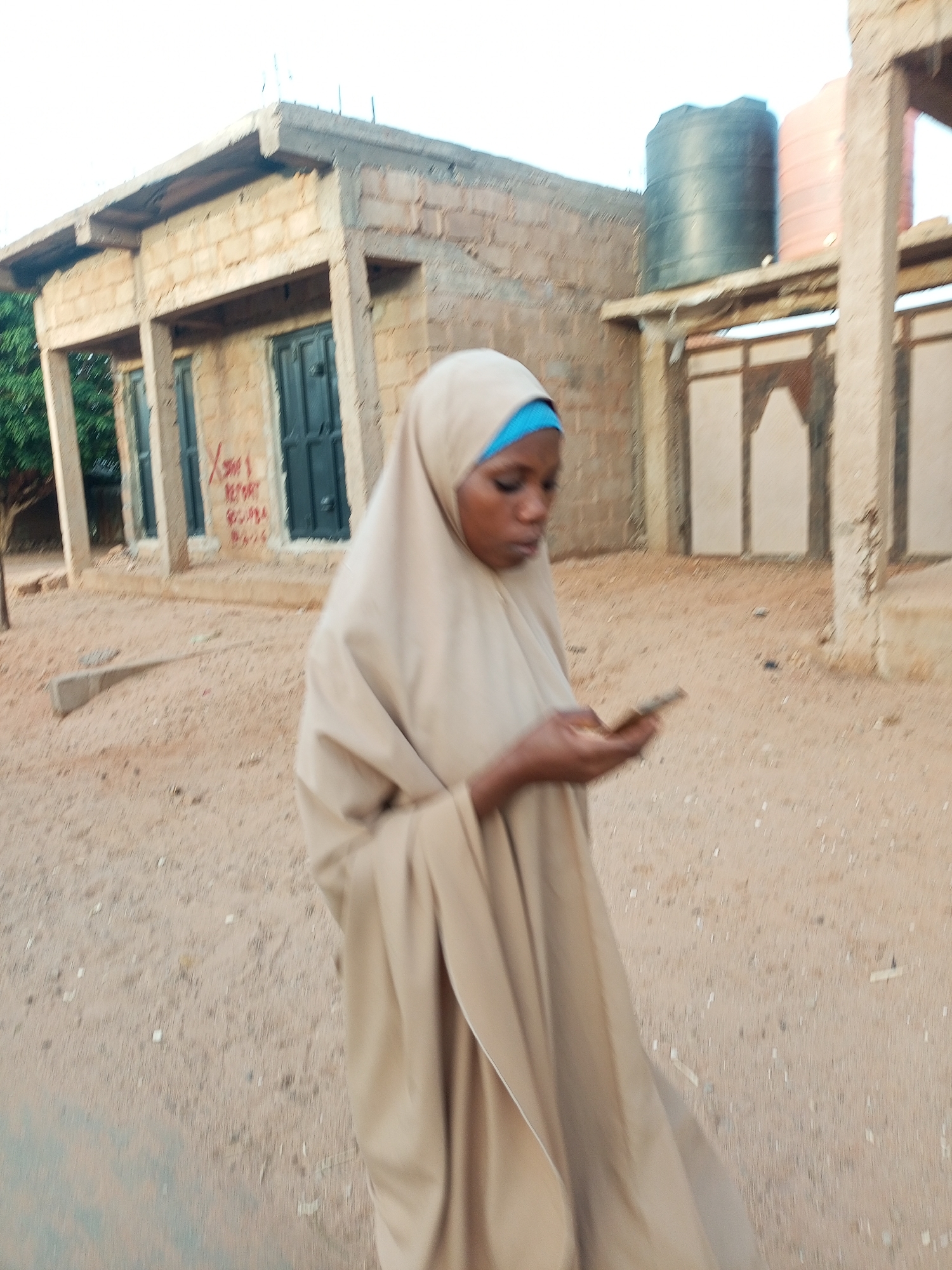 A woman in a tan hijab and blue headband is standing on a dirt road, looking at a cell phone in her hands. In the background is a partially constructed building with dark doors and what appears to be red graffiti on the wall. Behind that building are two water tanks, one black and one tan. The location is Tempure, Nigeria.