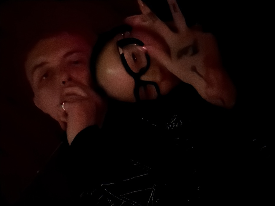 Two individuals pose in near darkness. One on the left has short hair and appears to be touching their face. The other wears glasses and makes a hand gesture with their fingernails visible. They are dressed in dark clothing, potentially with embroidered patterns. The overall image quality is low due to the limited light, obscuring details.