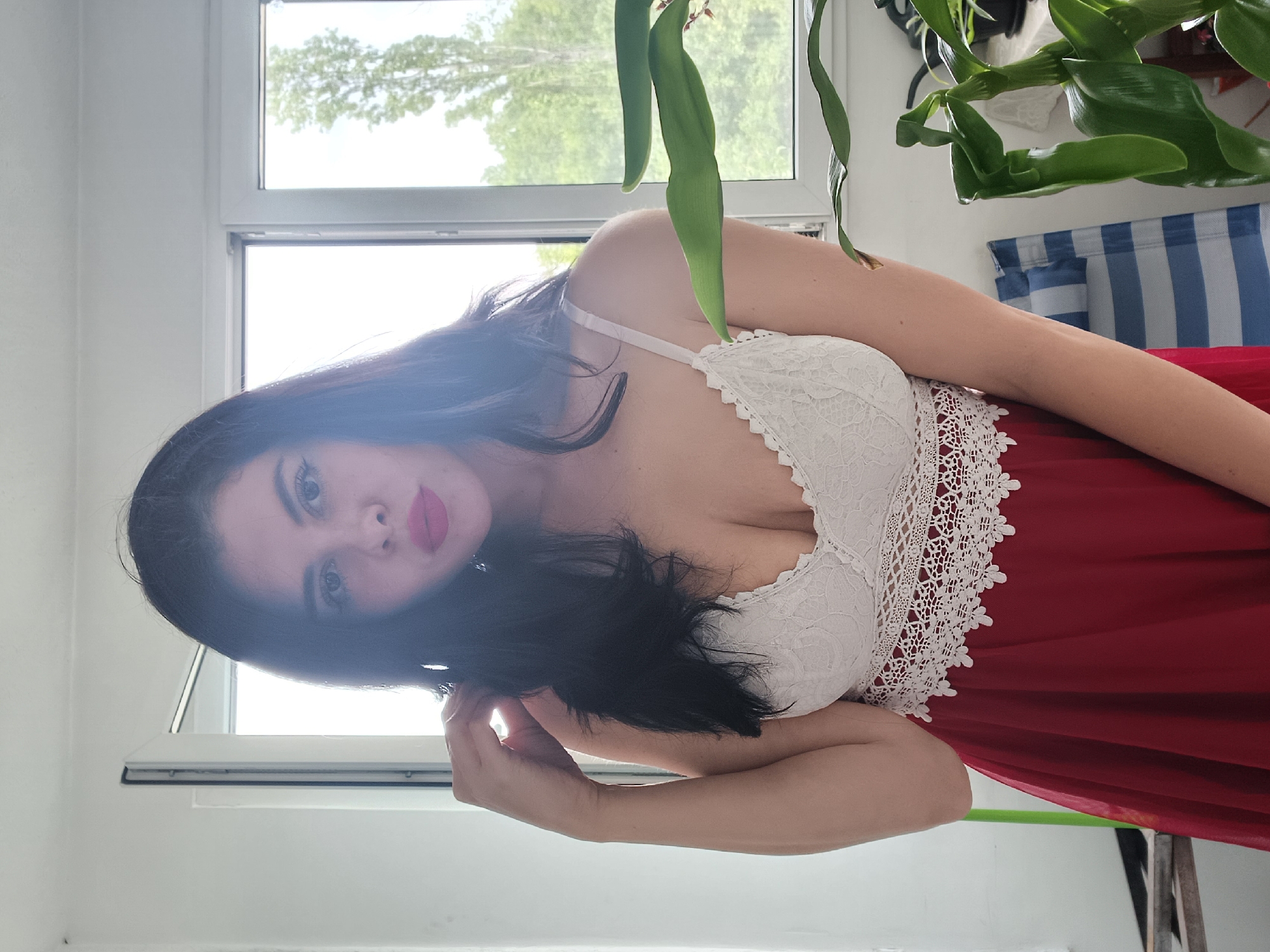 A young woman poses indoors in front of a window, likely in an apartment or home. She has dark hair and fair skin, wearing a white lace bralette and a red dress or skirt. Her hand is near her hair, suggesting a pose. Behind her is a window with an outdoor view, possibly showing trees. To the upper right, there is a plant with broad green leaves hanging from the top of the frame. A striped blue and white fabric (possibly a curtain or blanket) is also visible. The background suggests an interior setting, likely in a residential area. Without additional information, it is impossible to verify the exact location in Sofia, Bulgaria, as it is only specified to be the city in general.