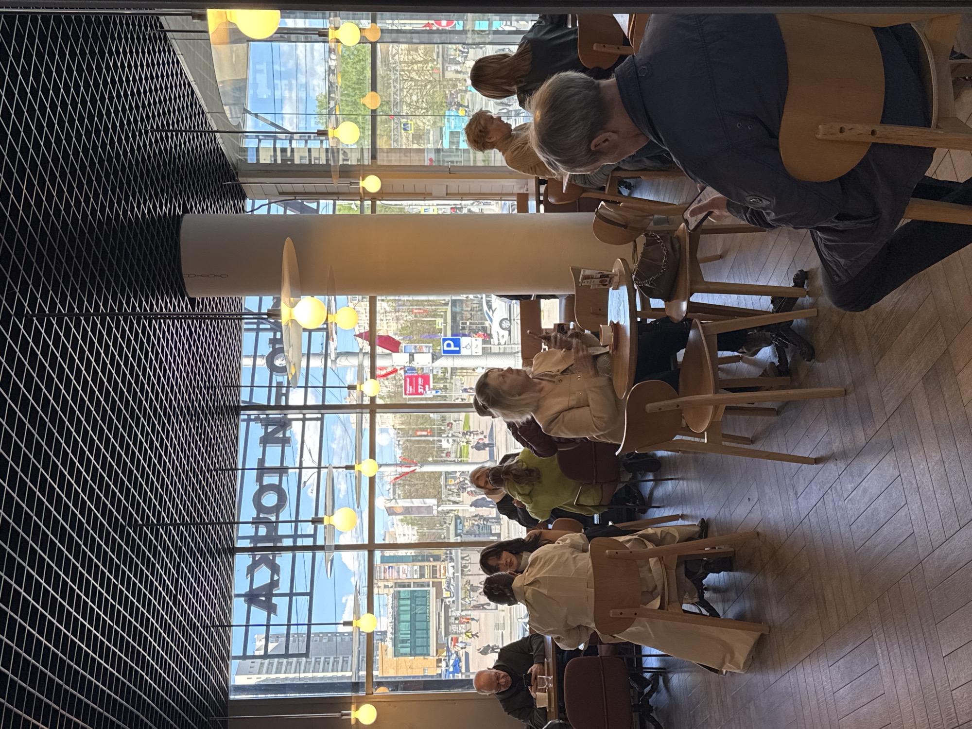 A photograph taken from inside a cafe in Saint Petersburg, Russia, shows several people seated at tables. The cafe has large windows providing a view of a city street with pedestrian traffic and buildings. Light fixtures consisting of multiple globe lights are suspended from the ceiling. A large, cylindrical structural column is visible within the cafe's interior. People are sitting at tables in groups, appearing to be eating, drinking, and talking. A man at the nearest table is looking at a device, possibly a phone. The cafe has a herringbone pattern wood floor. The exterior cityscape includes signage and a blue parking sign.