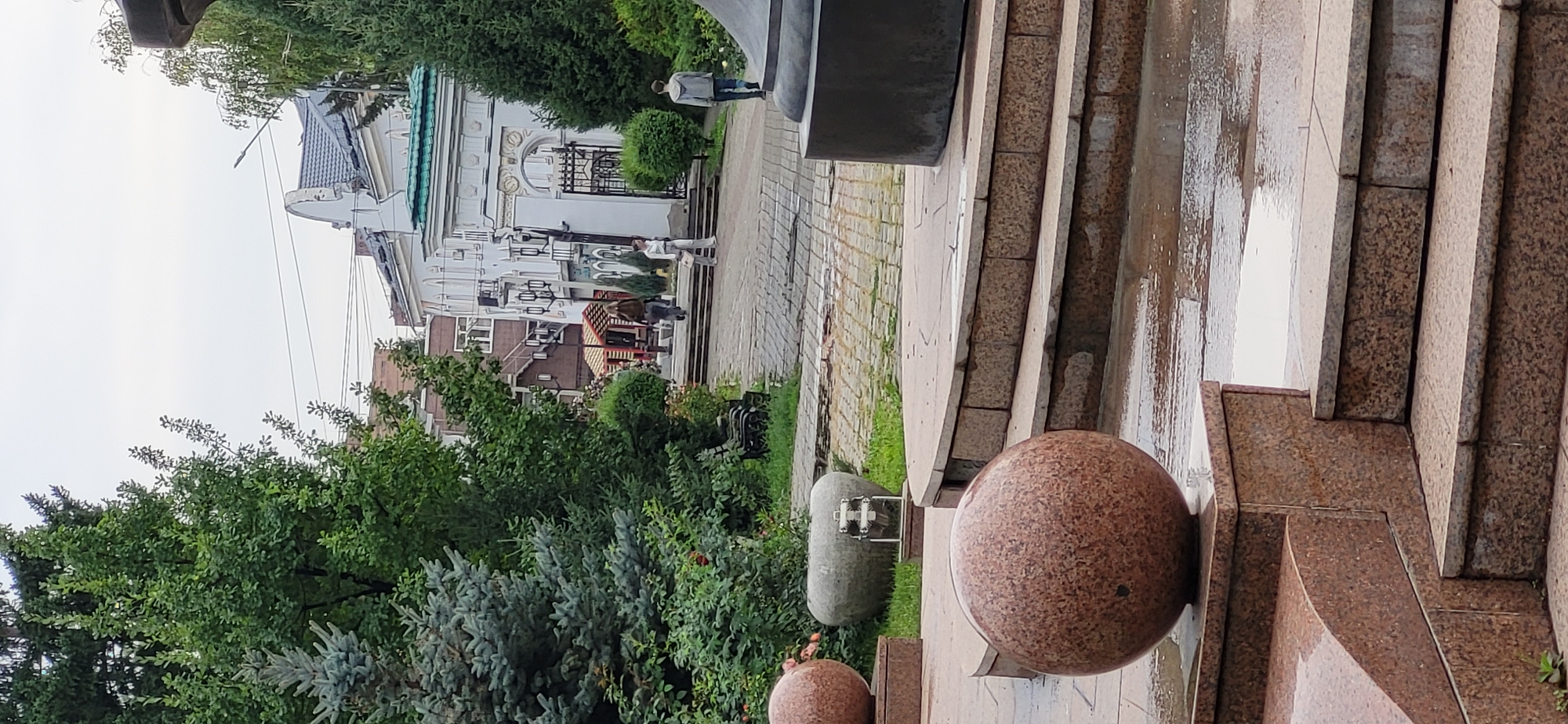 This image depicts an outdoor scene in Oskemen, Kazakhstan, likely within a park or public square area. In the foreground, there are decorative granite spheres and tiered steps, some of which appear wet, suggesting recent rain or a water feature. Lush green trees and shrubs frame the scene. In the mid-ground, a paved walkway leads towards a white building with an ornate entrance featuring a metal gate and arched structures. Several people are visible walking on the path and near the building. The architecture of the building suggests it might be a historical or significant structure. The overall atmosphere is serene, with a mix of natural elements and urban design.