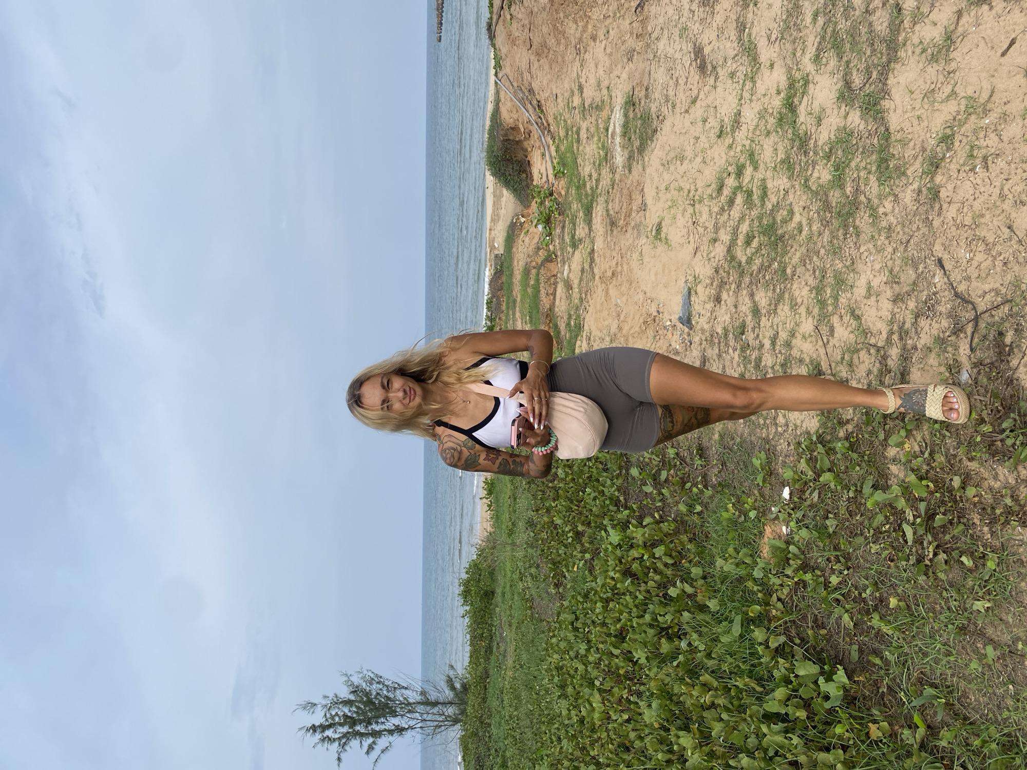 A young woman stands outdoors on a sandy path adjacent to a beach and the ocean. She is positioned centrally, facing slightly towards the camera with a soft smile. She has long blonde hair and visible tattoos on both arms. She is dressed in a white sports bra with black trim and grey high-waisted shorts, complemented by light-colored woven sandals. She holds a light pink or beige soft pouch bag in her left hand and appears to have a phone or similar item.
The setting is a coastal landscape. In the foreground and midground, the ground is sandy with scattered patches of green vegetation, including grasses and broad-leafed plants. A small, sparse tree is visible towards the lower left. To the right, the sandy terrain appears to form a gentle slope or bank leading down to the water's edge. The ocean stretches across the background, displaying muted blue-grey waters with subtle waves near the shore. A distant, dark structure, possibly a pier or breakwater, extends into the sea on the far left. Above the horizon, the sky is a pale blue with very faint cloud formations.
The image captures a relaxed moment during the daytime, under bright, clear weather. There is no visible text in the image. The scene suggests a tranquil seaside location, consistent with Tien Thanh Ward, Vietnam.