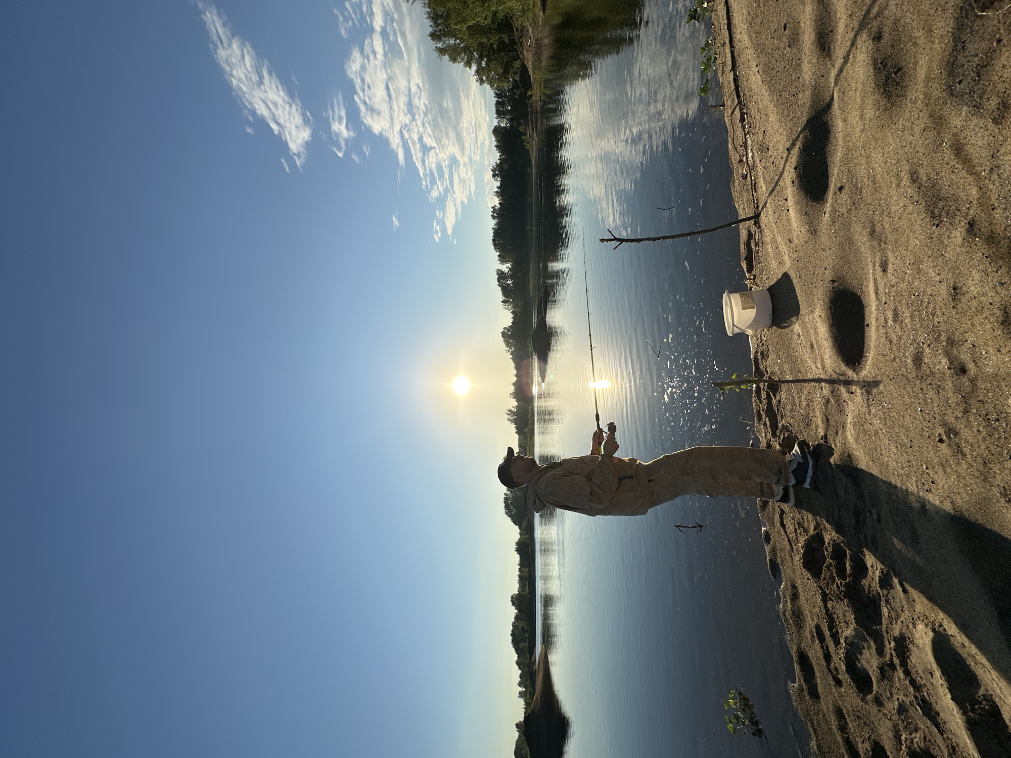 An adult male, dressed in camouflage fishing attire and a baseball cap, stands on a sandy bank by a calm body of water, casting a fishing rod. The sun, low in the sky, casts a warm glow across the scene, reflecting on the water and creating long shadows on the sand. The background features a treeline on the opposite bank, with a clear blue sky dotted with a few wispy clouds. A white bucket and a thin stick are placed on the sand near the fisherman. The image captures a serene moment of recreational fishing, likely in a natural setting near Noviy, Russia, given the foliage and waterbody visible.