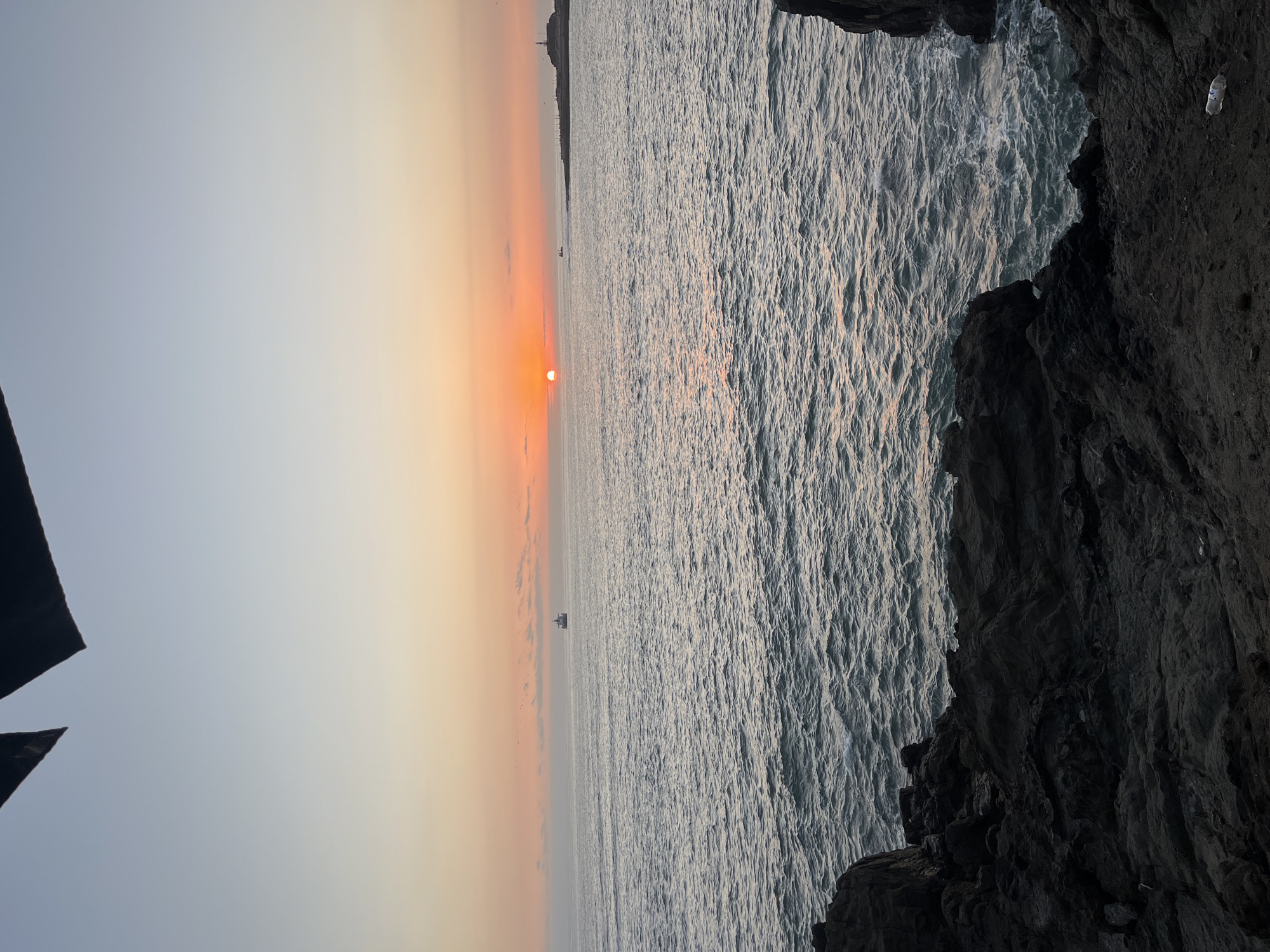 The image shows a sunset over the ocean, taken from a rocky cliff. The sun is partially obscured by a hazy horizon. A small boat is visible on the water. Arica, Chile.
