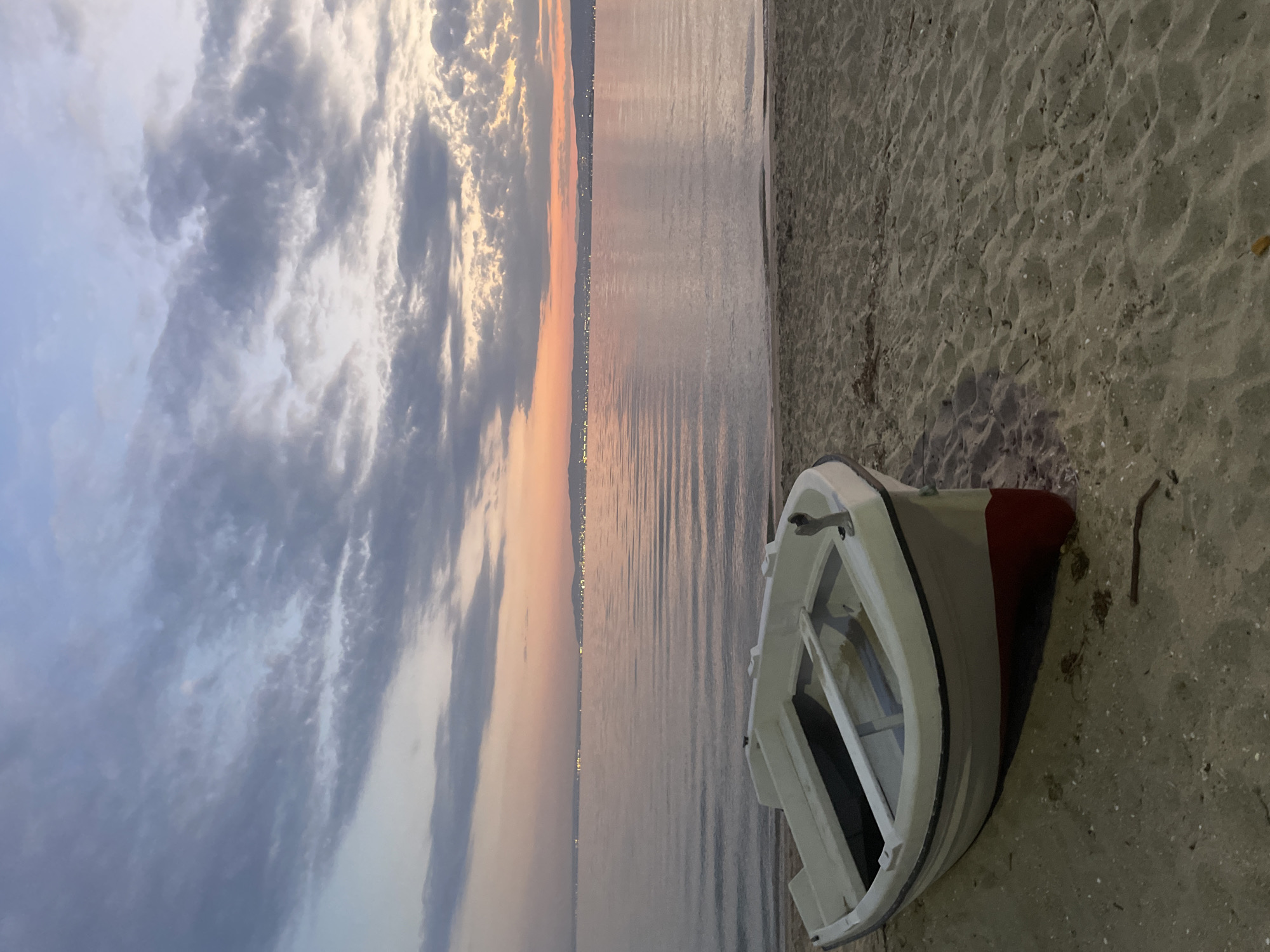 A white and red rowboat is beached on a sandy shore in Neoi Epivates, Greece, facing a calm body of water under a dusky sky. The water reflects the soft hues of the sunset, with a distant, illuminated shoreline visible across the expanse. The sky is filled with scattered clouds in shades of gray and pink, suggesting either dawn or dusk. The sand is textured with footprints and debris. No people are present in the scene.