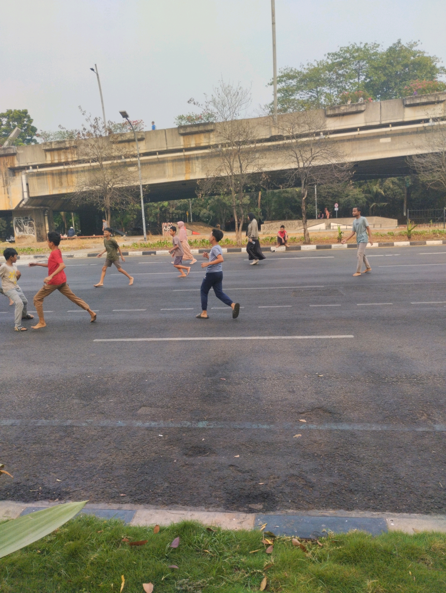 In Bekasi, Indonesia, a group of young people are engaged in activities on a paved road adjacent to a green area and a large overpass. The overpass, made of concrete, shows signs of aging with visible rust stains. Underneath the overpass, trees and foliage create a dense backdrop. Several individuals, predominantly children, are running and playing on the road, which has white lane markings. Some wear casual clothing like t-shirts and pants, while others are in more traditional attire. A few adults are also visible in the background, observing or walking. The foreground features a strip of lush green grass with fallen leaves, bordering the road. The overall scene suggests an outdoor recreational activity in an urban setting with significant infrastructure.