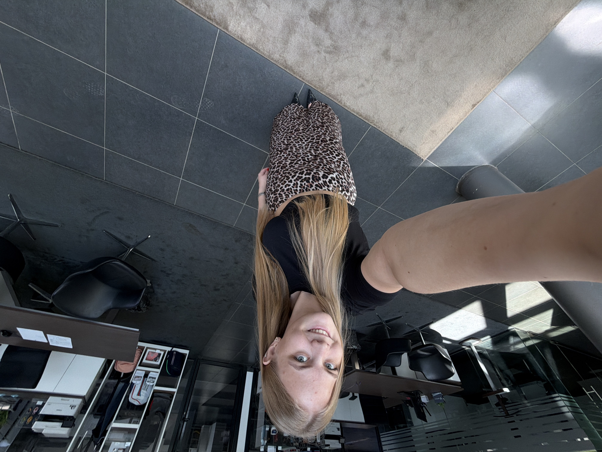 The image shows a blonde woman in a black top and leopard print skirt standing in an office space. She is facing the camera and appears to be taking a selfie, as her arm is visible extending to the side. The office space includes tiled flooring, a rug, desks, chairs, and filing cabinets. Outside the windows are visible buildings, suggesting an urban location. The location is not identifiable as Izhevsk, Russia, based on the visual information alone.