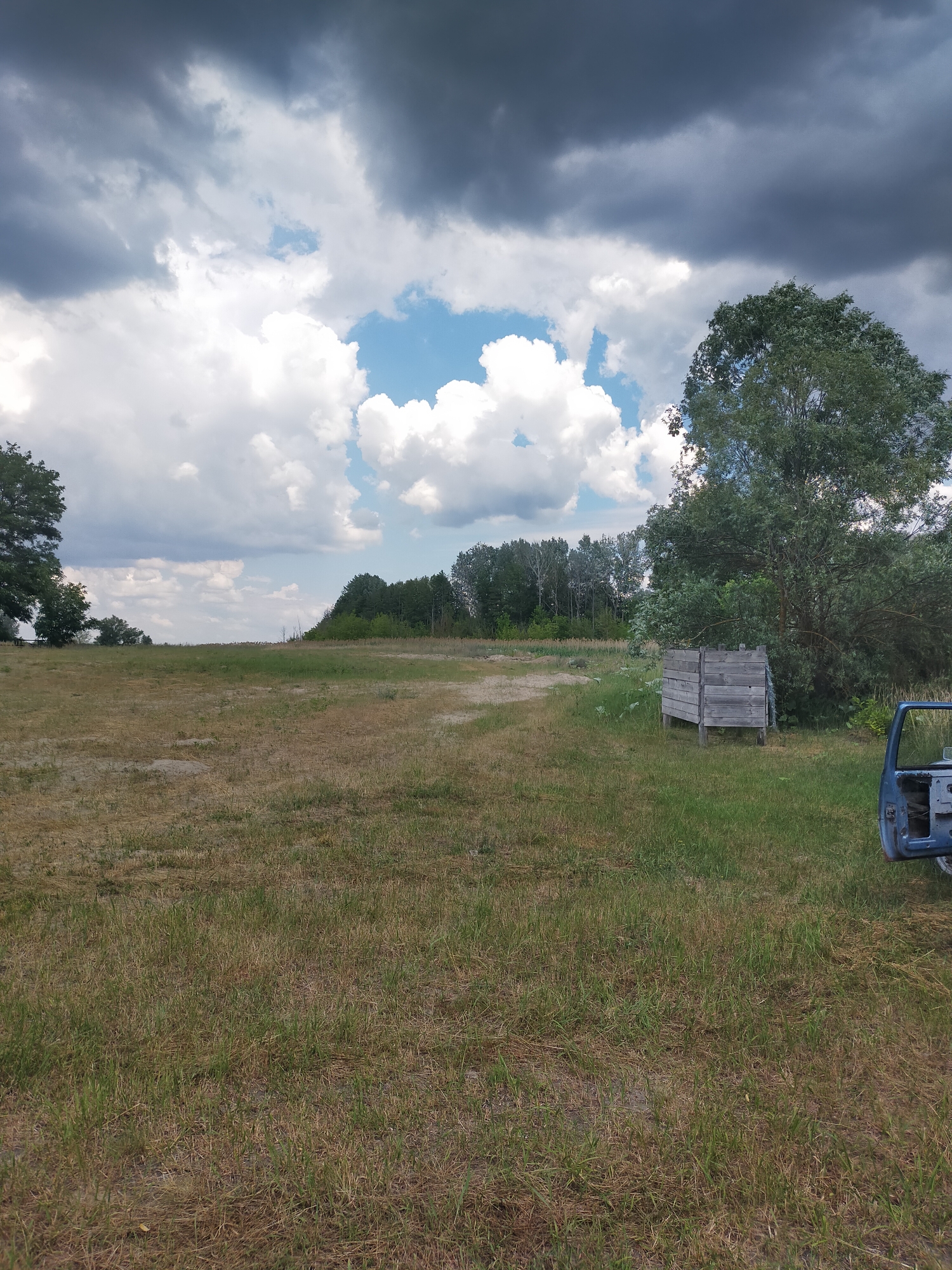 The image depicts an outdoor scene in Ustyvytsia, Ukraine, featuring a grassy field under a partly cloudy sky. The sky is dramatic, with dark storm clouds dominating the upper portion and bright white cumulus clouds interspersed with patches of blue sky. In the foreground, dry, yellowish-brown grass covers the ground, with areas of greener grass present. To the right of the frame, a weathered wooden structure, possibly a bin or barrier, stands in the grass. Adjacent to this structure, a blue car door, detached from a vehicle, is visible on the ground. In the background, a line of trees forms a dense forest, with a few lighter-colored trees mixed in. The terrain appears to be slightly elevated, sloping upwards towards the treeline in the distance.