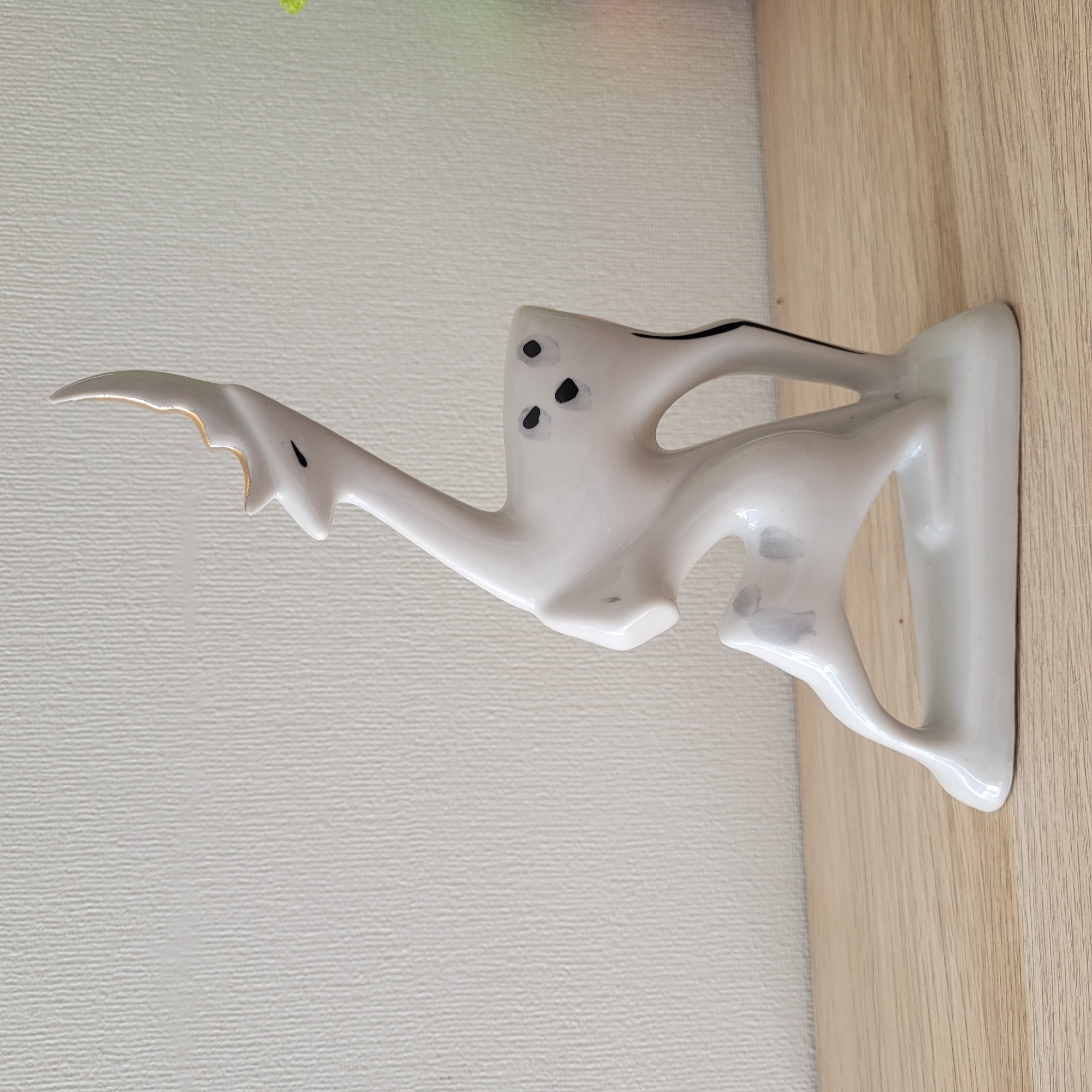 A white ceramic figurine of a stylized deer is placed on a light-colored wooden surface against a textured white wall. The deer is depicted in a dynamic pose, with its body contorted and its head raised. It has a single horn with a gold tip, small black eyes, and black spots on its body. There are also smudges of gray on the deer's legs. To the right of the figurine, a hint of green and red light is visible, suggesting the presence of another object or a colored light source outside the frame. The image appears to be taken indoors, possibly in a home or a shop in Jūrmala, Latvia, though no specific landmarks are visible. The lighting is soft and diffused, casting subtle shadows on the surface.