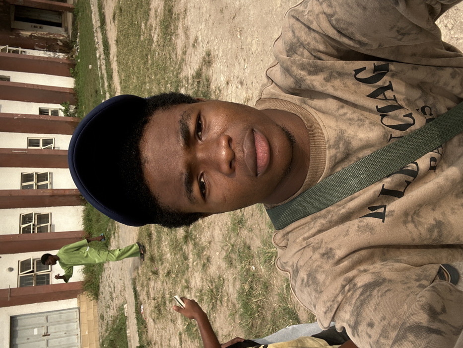 An outdoor daytime image, captured in Ihiagwa, Nigeria, features a young Black male in the foreground taking a selfie. He wears a dark blue baseball cap, a brownish-beige tie-dye sweatshirt with "BALENCIAG PARIS" text, and a dark green fabric shoulder strap crossing his chest. He maintains a neutral expression, looking directly into the camera. To his immediate left, a dark-skinned arm and hand are visible, holding a white smartphone horizontally. In the midground, a slim Black male, dressed in a light green long-sleeved tunic and matching trousers, walks away from the camera, turning his head slightly to the left. He appears to be holding a small blue object in his right hand and wears light-colored sandals. The ground is composed of light brown dirt with patches of sparse green grass. The background reveals a multi-story building with light-colored walls, prominent vertical brown architectural elements, and multiple open rectangular windows with light frames.