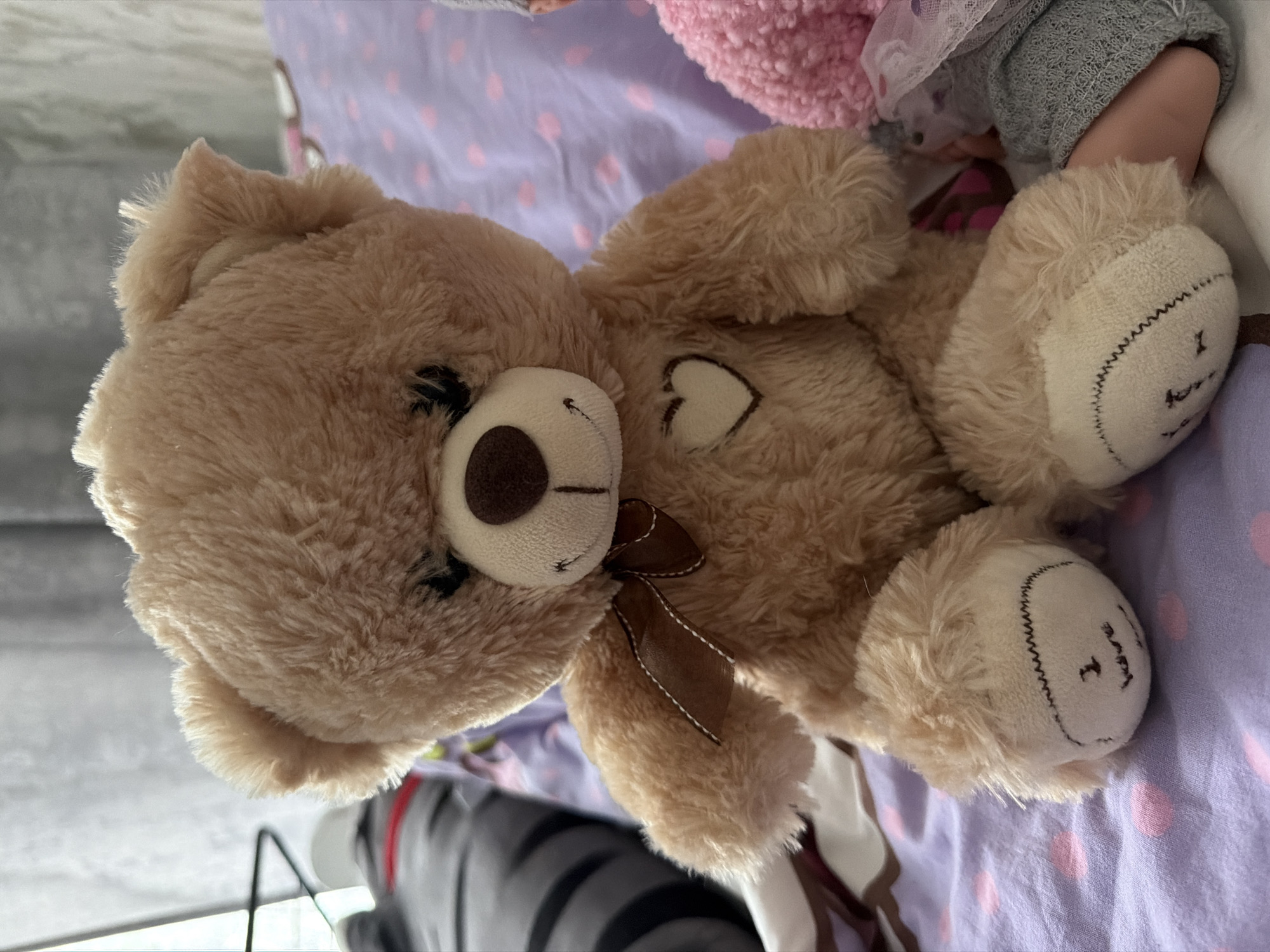 A tan teddy bear lies on a lavender sheet with pink polka dots. The bear has a brown nose and a brown bow. There is a small white heart on its chest. The bear's feet have white pads with black stitched smiles.
The location is not apparent from the image. The city of Noviy, Russia is not apparent from the image.