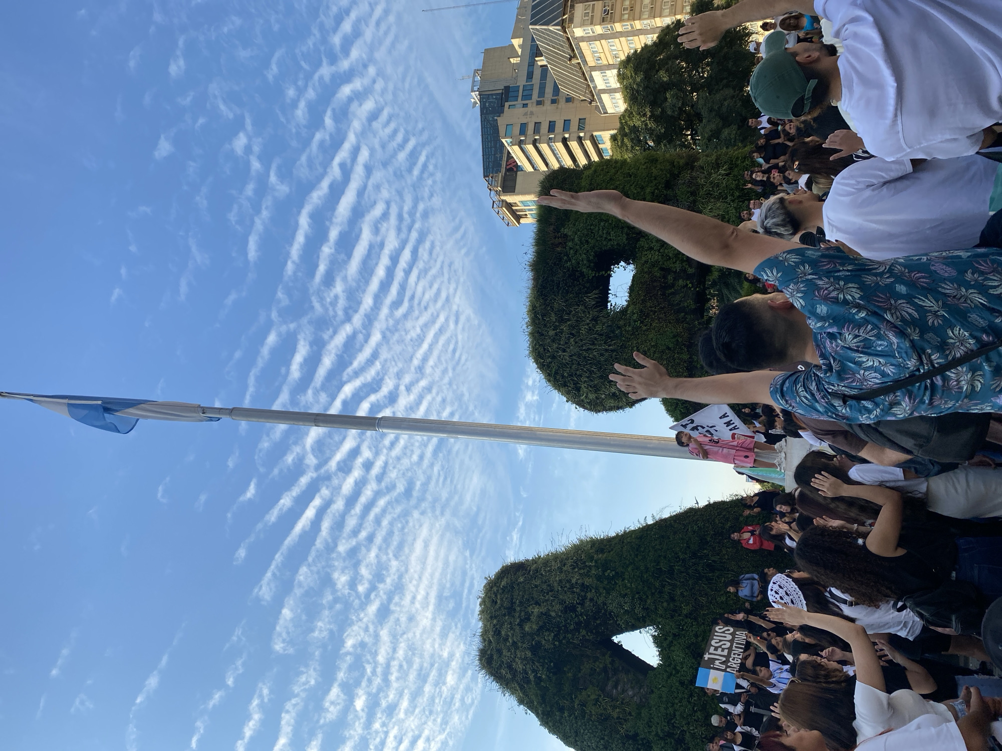 The image captures a scene in Buenos Aires, Argentina, likely at Plaza de la República near the iconic Obelisco landmark, which is partially visible in the background. A large gathering of people is present, many with their hands raised. An Argentine flag is flying on a pole nearby. The scene also includes topiary sculptures. A sign bearing the words "Jesus Argentina" can be seen within the crowd. The sky is bright blue with patterned clouds. The image appears to be taken from a low angle, looking upwards towards the sky and the crowd.