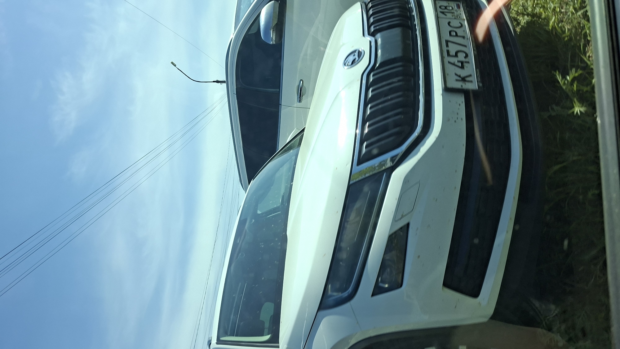 The image shows the front of a white Škoda Kodiaq car. The license plate reads "K 457 PC 18 RUS," indicating registration in Russia, specifically the 18th region, which corresponds to the Udmurt Republic. The photograph appears to have been taken from inside another vehicle, with the Škoda Kodiaq parked or stopped alongside. The background shows a blue sky, power lines, and some greenery, possibly roadside vegetation. Based on the license plate, the scene is likely in or around the city of Прикамский, Russia, which is located in the Udmurt Republic.