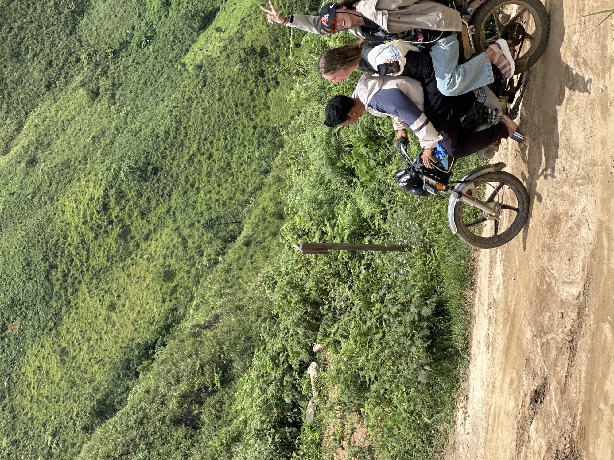 In Ta Xua, Vietnam, three individuals ride on a motorcycle along a dirt road. The person in the front is driving, wearing a blue and white jacket and black pants. Behind them, a woman in a white t-shirt and jeans holds a smartphone. The person in the back, wearing a beige jacket and a black baseball cap, raises their hand in a peace sign. The motorcycle is a mid-sized model with visible suspension and wheels. The environment is mountainous and lush, with dense green vegetation covering the slopes of the hills. The road is unpaved and appears to be a trail, with tire tracks visible in the dirt. The lighting suggests it is daytime, likely under a clear or partially cloudy sky.