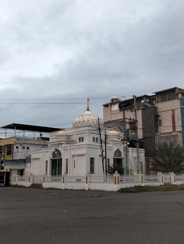A white mosque with a golden dome stands at a street corner under a cloudy sky. The building is surrounded by a low white fence with gold accents. Adjacent buildings are visible in the background, showing a mix of residential and commercial architecture. Power lines crisscross above the mosque, adding a modern element to the otherwise classic structure. The overall atmosphere is serene despite the urban setting.