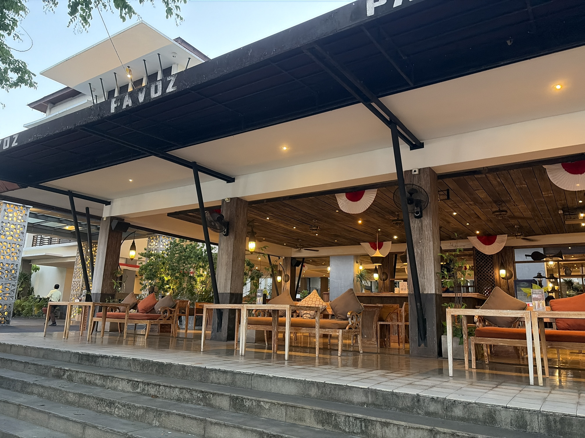 The image depicts the outdoor seating area of a restaurant or cafe in Kuta, Indonesia, located on Jalan Kartika Plaza. The establishment, identified by the sign "PAVOZ" on the building's facade, features a covered patio with wooden tables and chairs arranged on a tiled floor. Several wooden sofas with orange and brown cushions are visible. The ceiling of the patio is made of wood and adorned with decorative red and white fans. Lighting fixtures hang from the ceiling. Green foliage and trees are present in the background. To the left, a person in a light-colored shirt is walking. The area is accessible via a wide set of concrete stairs in the foreground. The sky is overcast, suggesting it might be early morning or late afternoon.