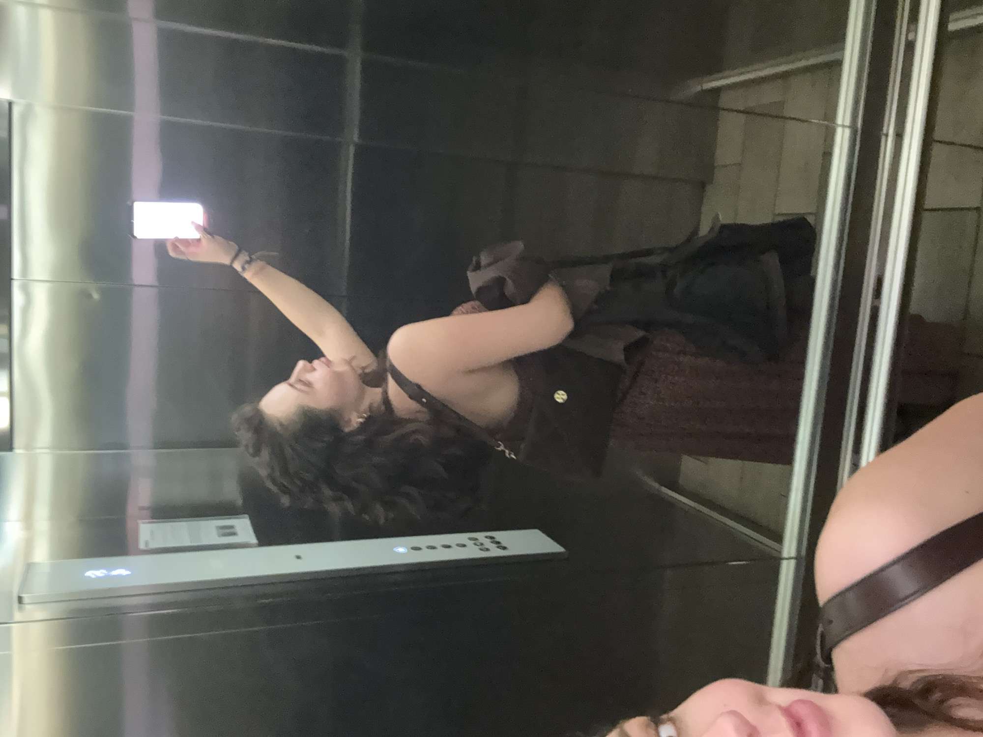 A woman is taking a selfie in an elevator. She is holding up her phone with her right hand. She has curly dark hair and is wearing a dark top with a cross-body bag. The elevator is lined with stainless steel panels. There are elevator control buttons visible. The reflection of another person is seen at the bottom of the image. The location is potentially Mexico City, Mexico, based on assumptions.