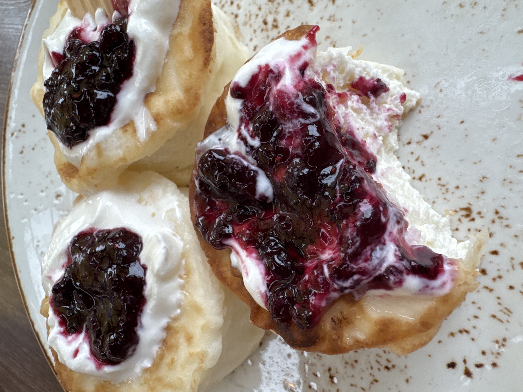 A close-up, high-angle shot captures a plate of traditional pancakes (likely *syrniki* or *oladi*) generously topped with a dollop of white cream and a dark berry jam or compote. There are at least three pancakes visible, with one in the foreground showing a bite taken out, suggesting it is actively being consumed.
The pancakes themselves are golden-brown and appear thick and fluffy. Each is crowned with a substantial portion of creamy white topping, which could be sour cream, yogurt, or whipped cream. Over this white topping, a rich, dark purple-red berry jam is spooned, featuring visible whole or chunky pieces of berries, possibly blueberries or blackcurrants, with glossy, sticky textures. The jam has bled slightly into the cream, creating an appetizing marbling effect, particularly on the partially eaten pancake where the jam is spread more broadly.
The food is presented on a light-colored, possibly ceramic, plate with subtle, speckled brown or rust-colored patterns, adding a rustic aesthetic. The lighting suggests an indoor setting, likely during breakfast or dessert time. There is no visible text in the image. The scene is set in Bishkek City, Kyrgyzstan.