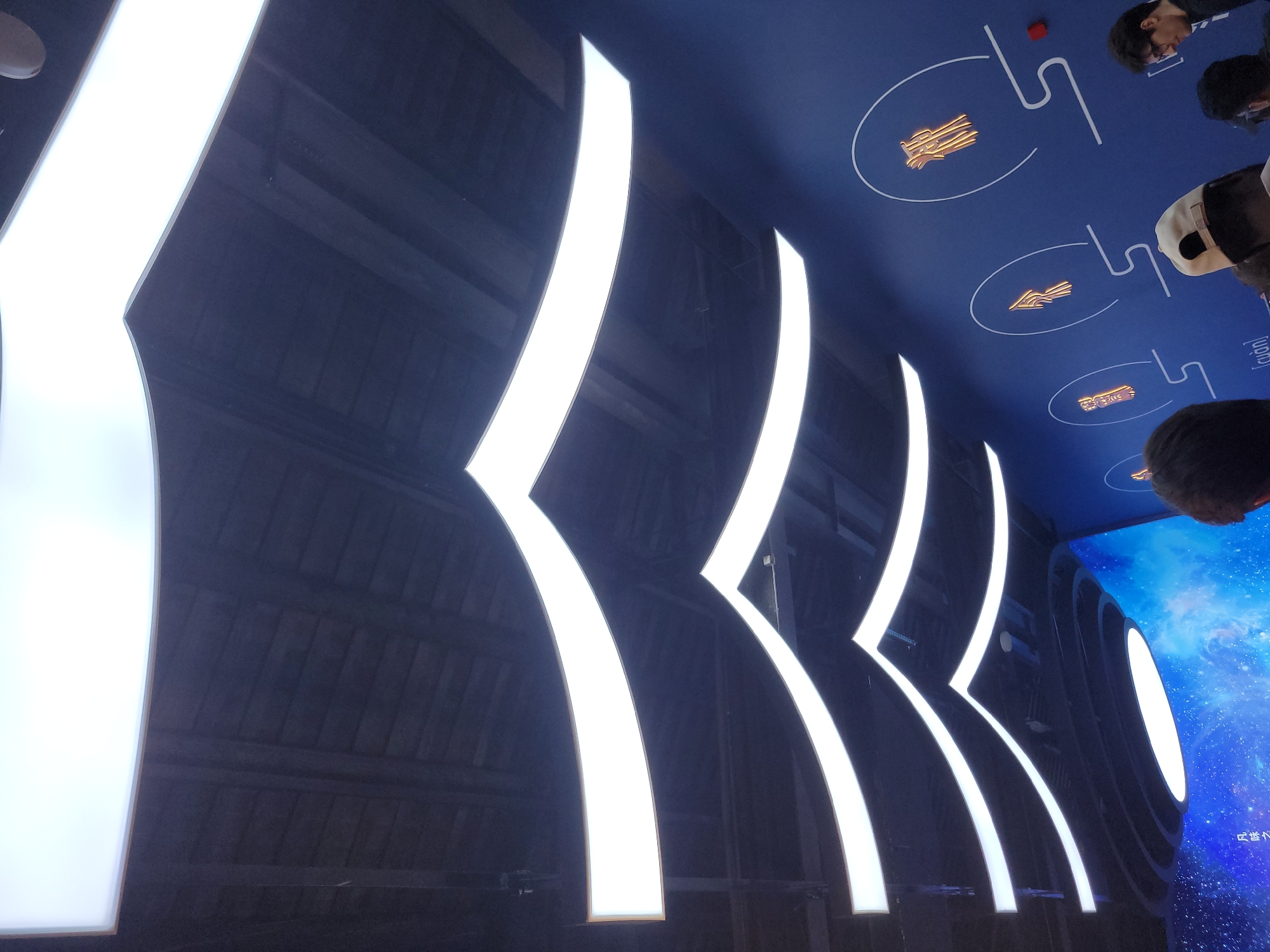 The image shows an interior architectural feature with repeating curved illuminated panels in the city of Hechuan, China. The panels are white and set against a dark blue or black surface. At the top of the image, several people are partially visible, looking down at the ceiling. There are depictions of vehicles on the ceiling.