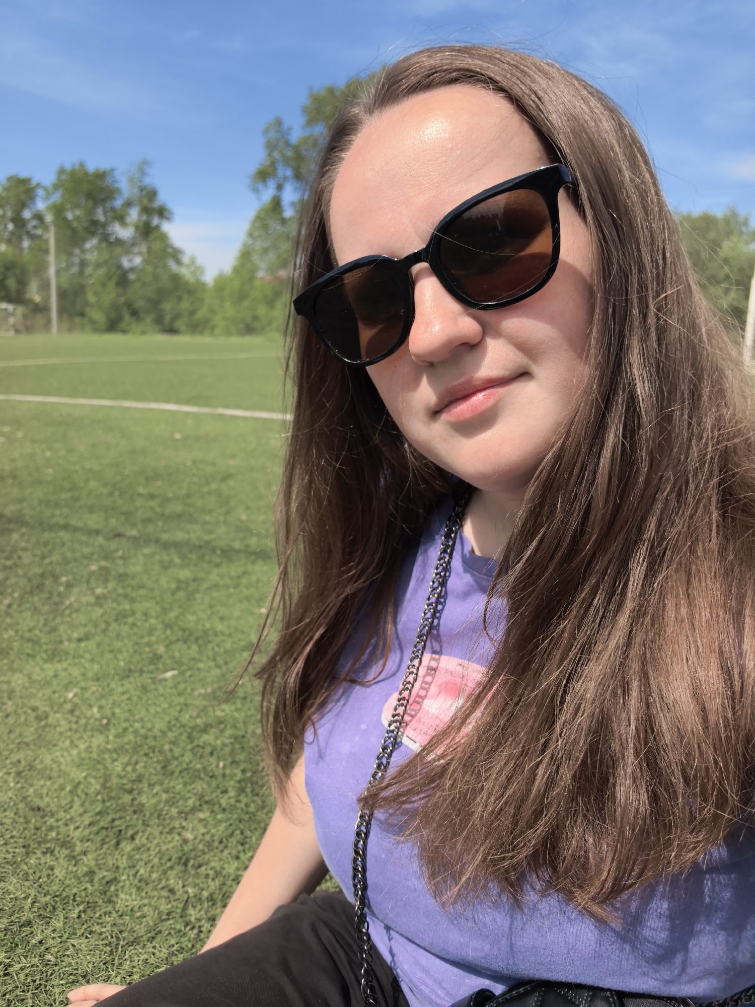 A young woman with long brown hair and sunglasses sits outdoors in Фролы, Russia, likely on a grassy field, as evidenced by the green turf, white lines, and soccer goal in the background. She is wearing a light purple t-shirt with a graphic and a silver chain necklace. The sky is blue with scattered clouds.