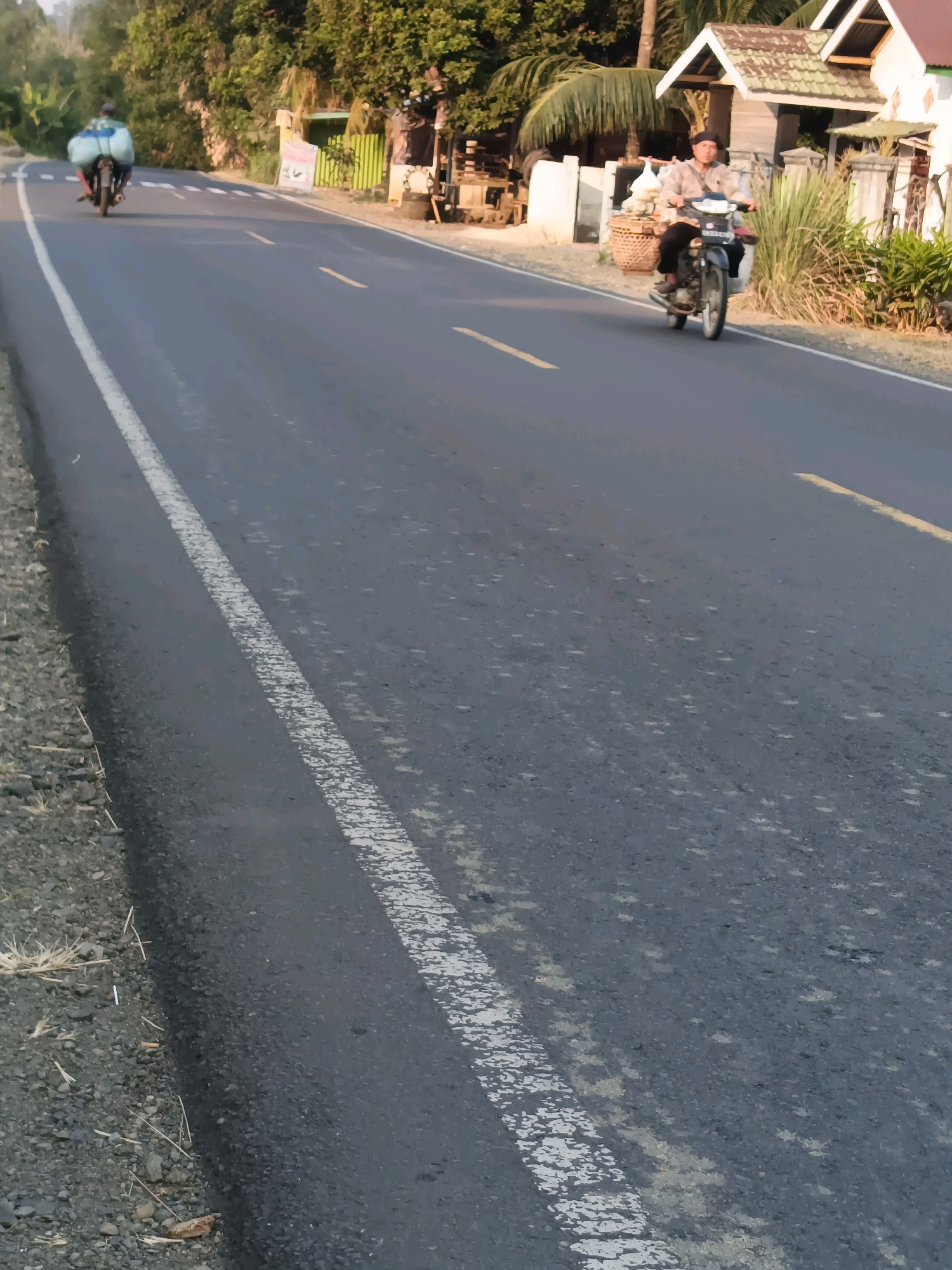 An outdoor, eye-level, medium shot captures a paved road with a white line marking the edge and a yellow line in the center, situated in Tanjung Sanai I, Indonesia. On the road, two people are riding motorcycles. In the foreground, a person in a light-colored collared shirt and dark pants rides a black motorcycle from the right side of the frame toward the left. They wear a dark cap and have a basket on the left side of their motorcycle. Further down the road, in the background, another person rides a motorcycle away from the camera, carrying a large, light-blue bundle on the back. The road is flanked by a rocky shoulder on the left and a grassy area with some residential structures and trees on the right. The lighting suggests it is daytime, likely late afternoon due to the warm tones.