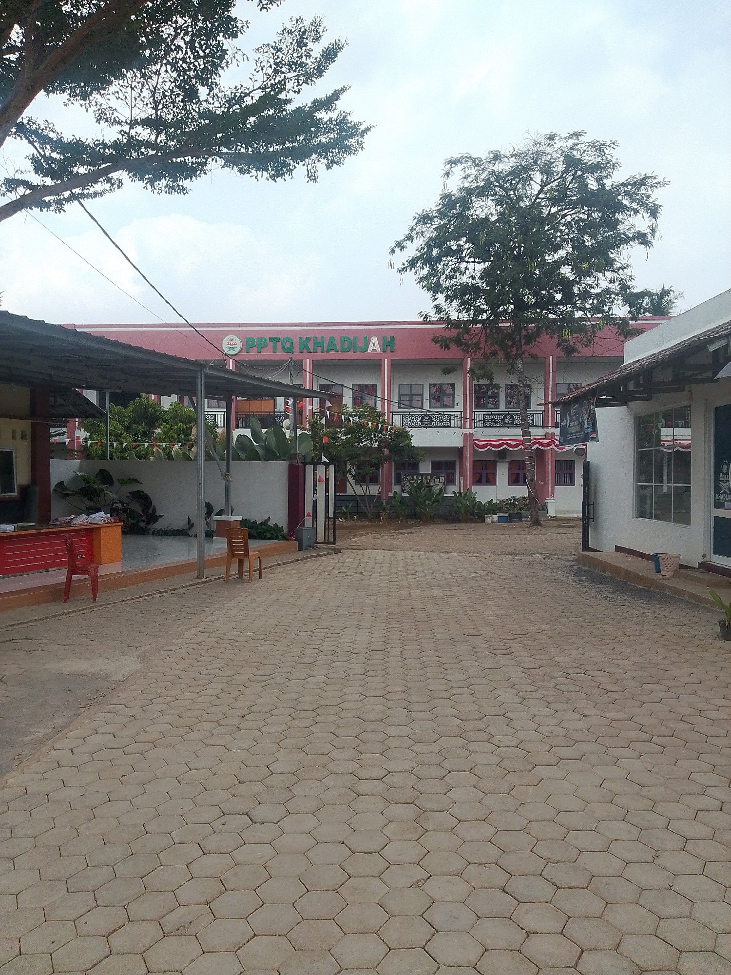 The image shows a courtyard paved with hexagonal concrete blocks. In the background, a two-story building with a red facade and white trim is visible. The building has multiple windows and decorative elements. A sign above the entrance reads "PPTQ KHADIJAH" in green letters. Trees are present on the left and in front of the building. A covered walkway runs along the front of the building, and there are tables and chairs in the courtyard. The scene appears to be an educational institution, possibly a pesantren (Islamic boarding school), in Kurungannyawa, Indonesia, given the name "KHADIJAH" which is often associated with Islamic foundations. The sky is overcast.