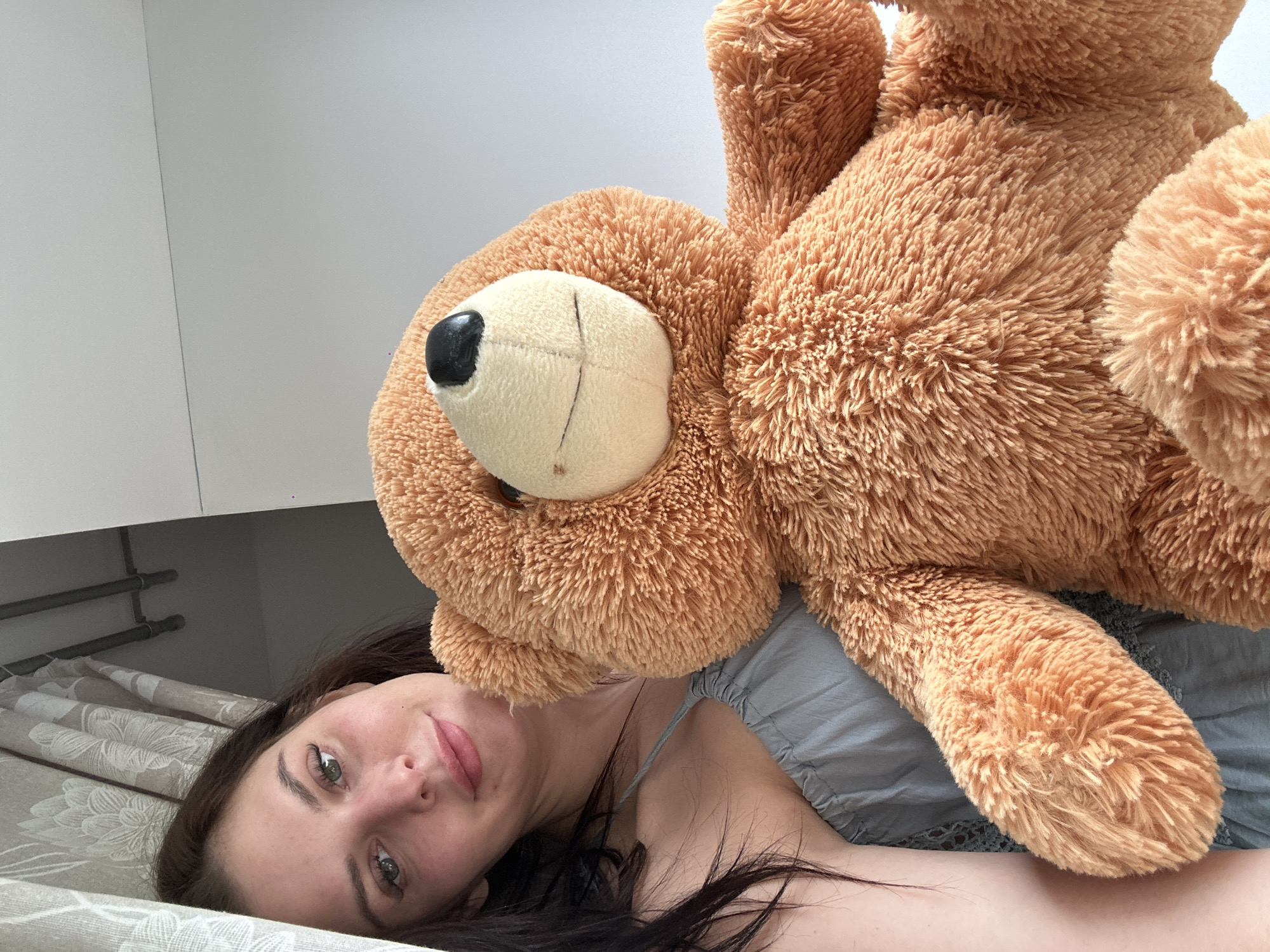 A woman with dark hair and green eyes smiles as she holds a large, fluffy, light brown teddy bear in front of her. She is wearing a light blue, sleeveless dress. She is positioned on the left side of the frame, and the teddy bear occupies the majority of the right side. The background appears to be indoors, with a light-colored wall and a window with floral curtains on the left.