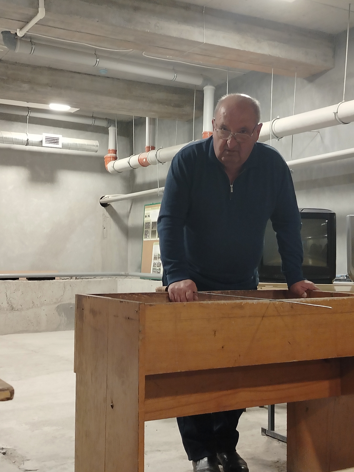 An older Caucasian male with glasses and a bald head is standing in what appears to be a basement or utility room. He is wearing a dark blue zip-up long-sleeved shirt, dark pants, and black shoes. He is leaning on a large, unfinished wooden structure, possibly a workbench or a piece of furniture in the process of being built or repaired. The man's hands are resting on the top edge of the wooden structure.
Behind the man, the environment includes exposed concrete walls and ceiling with visible pipes and ductwork running overhead. There is a poster or board on the wall. There is a small appliance visible. The space is illuminated by overhead lighting.
There is no information to place the image in Yerevan, Armenia.