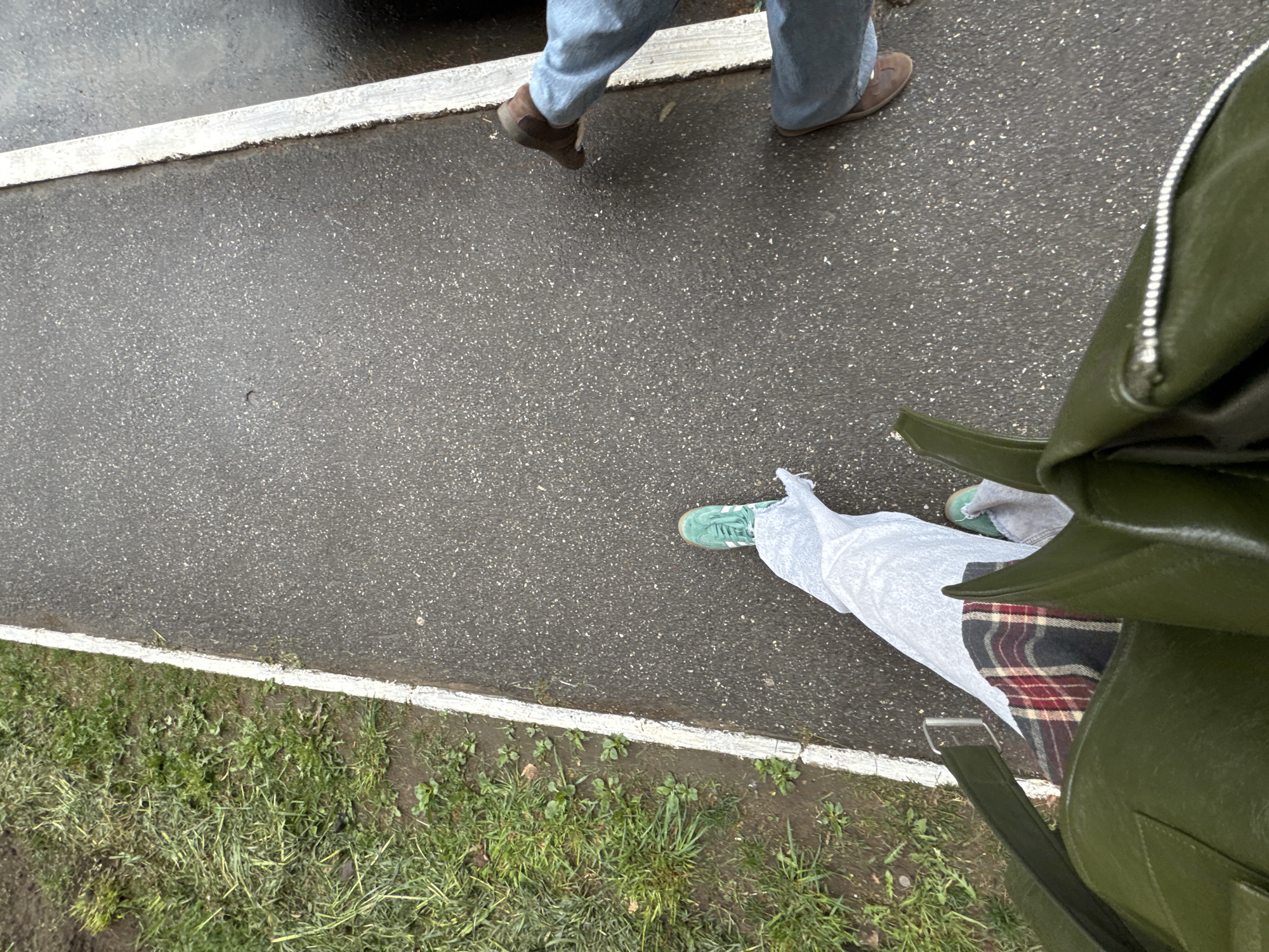 The image depicts a section of a paved area, likely a sidewalk or path in Chaikovsky, Russia, adjacent to a grassy area. A white line demarcates the paved surface. Two individuals are partially visible. One person is walking with their feet visible, wearing blue jeans and brown shoes. Another person is wearing light-colored pants, green sneakers, and a green bag on their shoulder. The bag contains a plaid item. The pavement appears wet.
