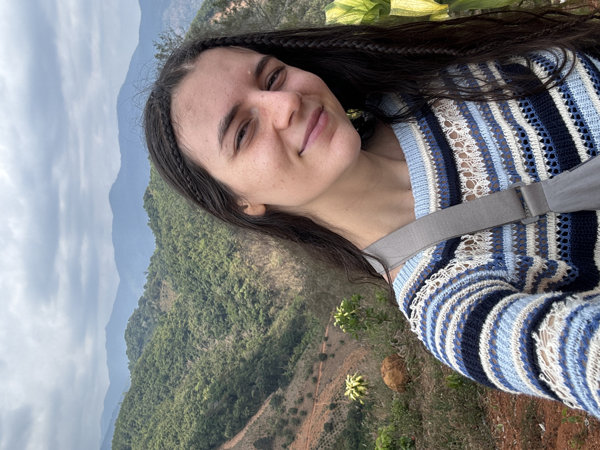A young woman is featured in a close-up selfie, smiling gently with closed lips and open eyes, looking towards the camera. She has long, dark hair, with small braids visible near her temples. She is wearing a distinctive, open-knit sweater patterned with horizontal stripes in light blue, white, and dark blue. A grey strap, possibly from a bag or harness, crosses diagonally over her chest.
The scene behind her is a vast, natural landscape, identified as Nao Sẻ, Vietnam. Rolling mountains, densely covered in green forest, stretch into the distance, appearing hazy and lighter blue under a soft, overcast sky. The immediate ground in the foreground and midground is composed of reddish-brown earth with scattered green vegetation and small plants.
The photo appears to have been taken during the daytime, likely under diffused light due to the cloudy or hazy weather conditions. The woman's expression conveys happiness and contentment, suggesting she is enjoying the scenic mountain views. There is no visible text in the image.