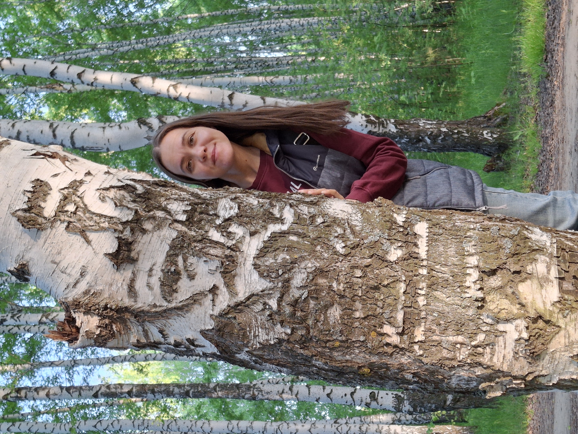 The image shows a woman leaning against a birch tree. She has long brown hair and is wearing a maroon sweater, a dark vest, and jeans. Her right arm is resting on the tree trunk, and she is smiling at the camera. The background consists of other birch trees, grass, and a dirt path. The image appears to have been taken in a wooded area or park, most likely in the city of Chaikovsky, Russia.