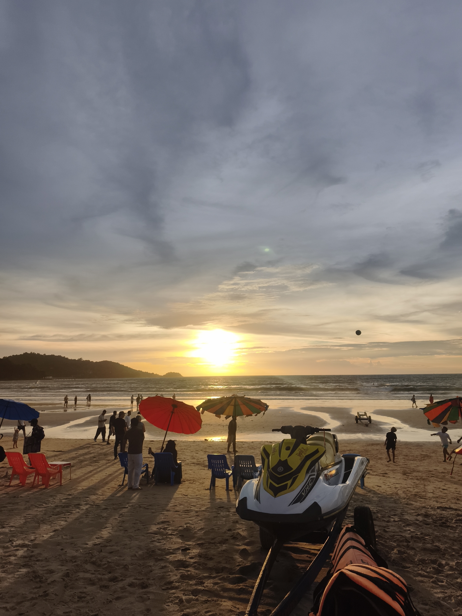 A beachfront scene unfolds at Patong Beach, Thailand, as dusk settles. The sun, a bright orb, hovers just above the horizon, casting a warm, golden glow across the sky and water. The sky is a dramatic canvas of grey and pale orange clouds, with a darker, more ominous layer overhead.
The sandy beach is populated by a scattering of people. Some are walking along the water's edge, their figures silhouetted against the receding waves. Others are clustered near colorful umbrellas, taking respite from the sun. Beach chairs, in bright red and blue, dot the sand, some occupied, others empty.
In the foreground, a white and yellow jet ski rests on a trailer, its sleek design ready for action. Its presence hints at the recreational activities available on this popular tourist spot. Further down the beach, a dark, spherical object hangs in the air, possibly a ball or a kite.
The ocean stretches out to the horizon, its surface rippled by gentle waves. The coastline is lined with lush green hills, their slopes disappearing into the hazy distance. The overall atmosphere is one of relaxed enjoyment, a typical evening at a bustling Thai beach.