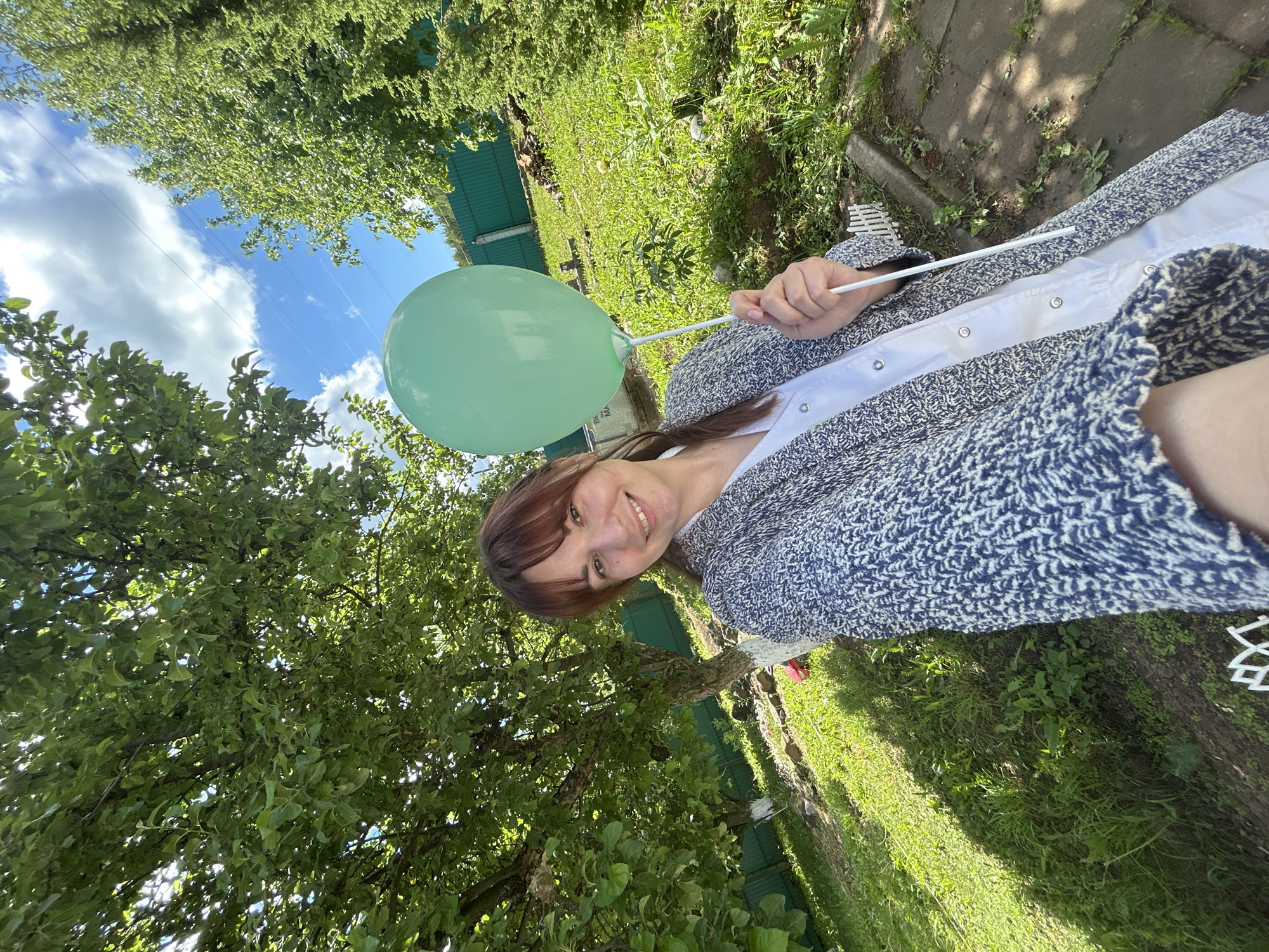 A woman in a gray and blue cardigan and white shirt is holding a green balloon on a stick. She is smiling and facing the camera. The background consists of green trees and grass, suggesting an outdoor setting. The sky with blue patches and white clouds is visible through the trees.