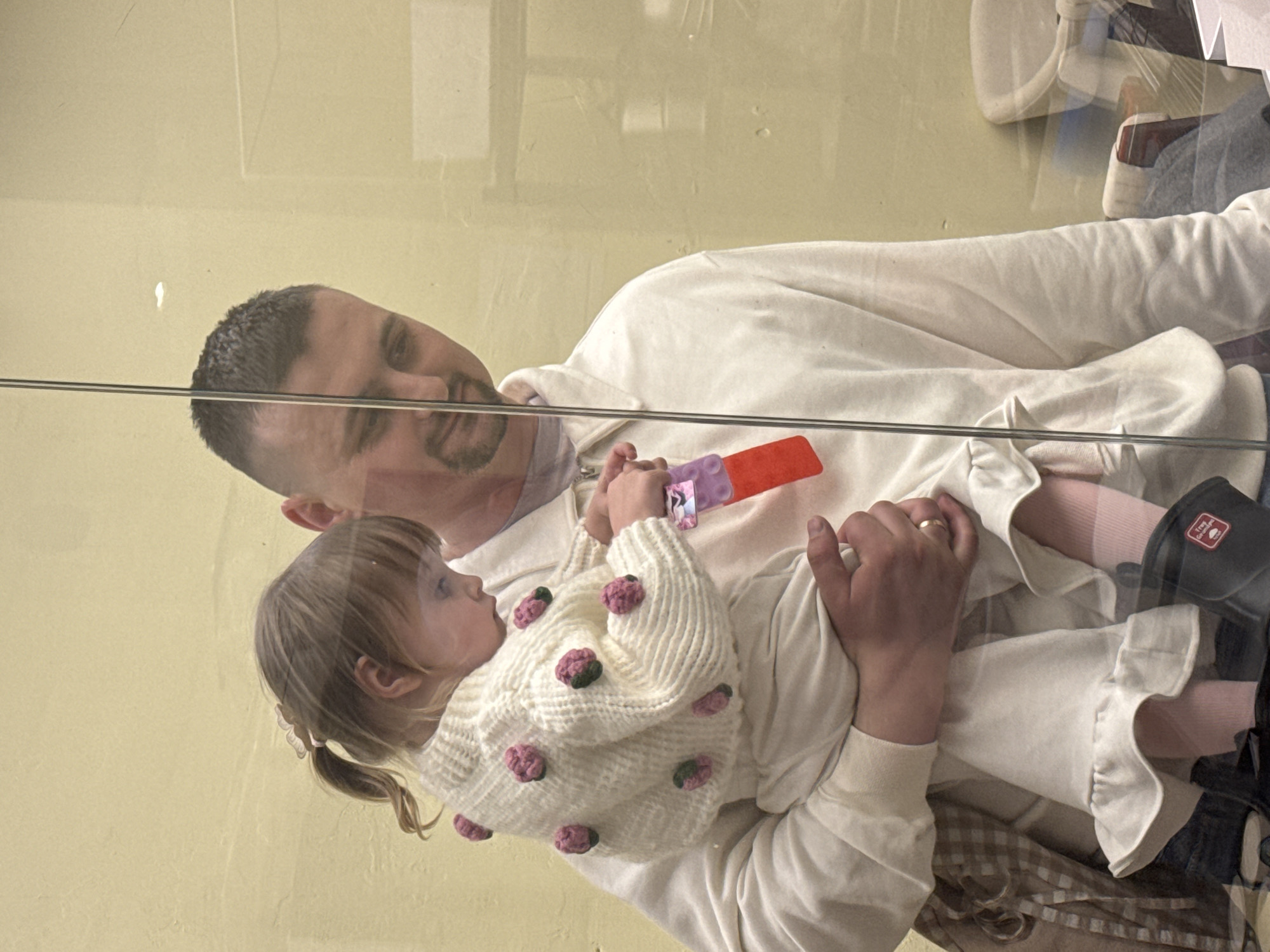 The image captures a tender interaction between a man and a young child, likely a girl, seen through a transparent barrier, possibly glass, in an indoor setting. The man, with short dark hair and a beard, is wearing a light-colored long-sleeved shirt and has a gentle smile as he looks down at the child. The child, with light brown hair pulled into pigtails, is dressed in a distinctive white knitted sweater adorned with numerous pink or red pom-poms. She holds a purple and orange/red toy and gazes up at the man.
A silver horizontal bar or railing spans across the image, reinforcing the presence of a transparent partition. The background consists of a plain, light yellow-beige wall, visible through reflections that also show faint outlines of other objects or furniture. The man wears a ring on his left ring finger. On the child's lower leg, a black object with white text is partially visible, which clearly reads "Young Care" in the top two lines, with further text below that is obscured. The scene evokes a sense of affection and connection between the two individuals. The location context is Khimki, Russia, and the indoor lighting suggests a soft, diffuse light source.