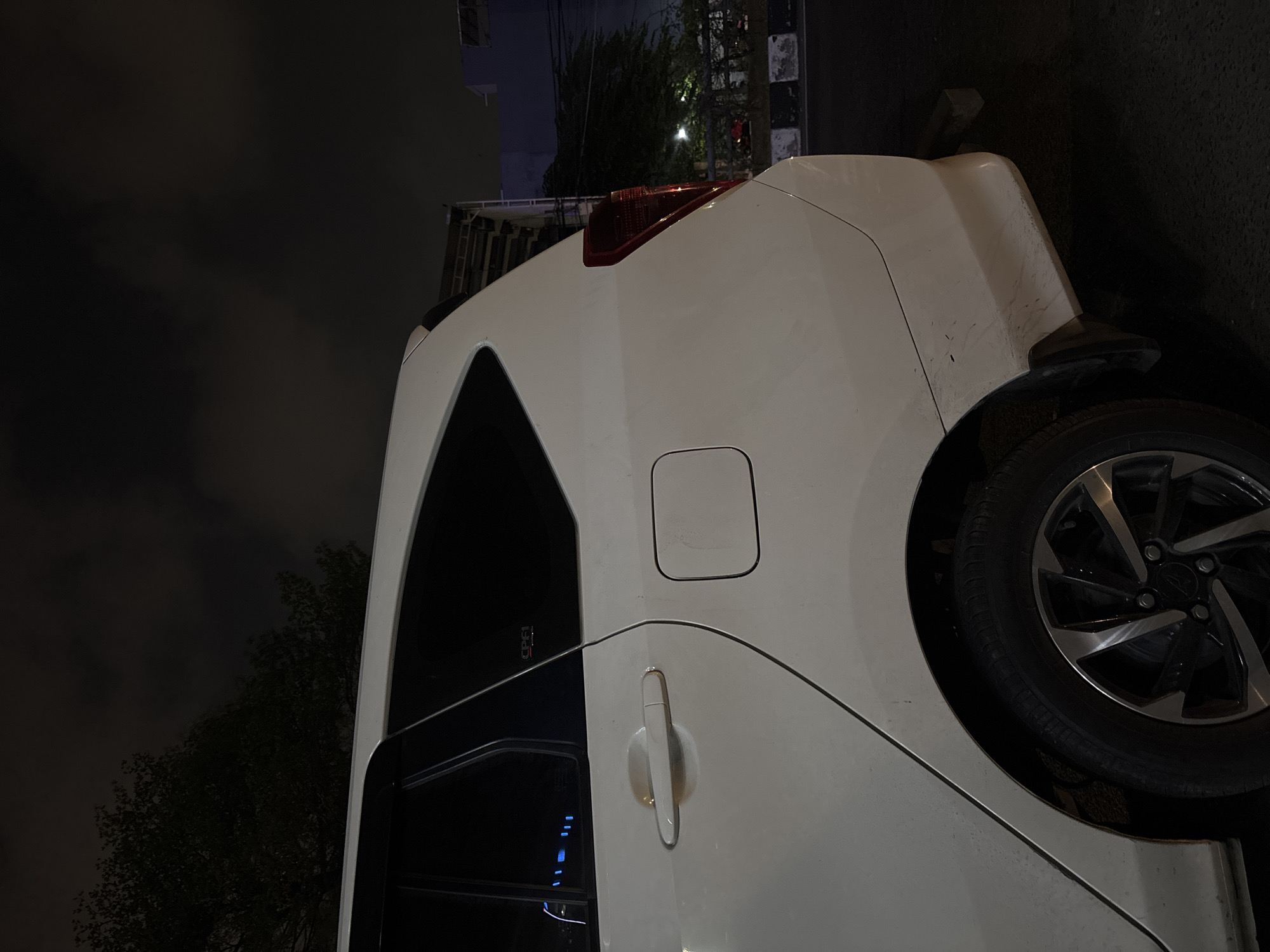 The image depicts the rear right side of a white car parked at night. The car is illuminated by ambient light, casting shadows. A tree with dark foliage is visible to the left of the car, and the night sky is overcast. In the background, there are structures, possibly buildings, and electrical wires. The scene appears to be on a street with a road surface and a curb. A portion of a truck or similar vehicle is partially visible behind the car. The car's taillight is red, and the wheels are silver and black alloy rims. The car has a fuel filler cap on the rear quarter panel and a door handle on the rear passenger door. A small emblem, possibly a car logo, is visible on the rear passenger window. The car appears to be slightly dusty. The environment suggests an urban setting in North Jakarta, Indonesia, given the time of night and the visible infrastructure.