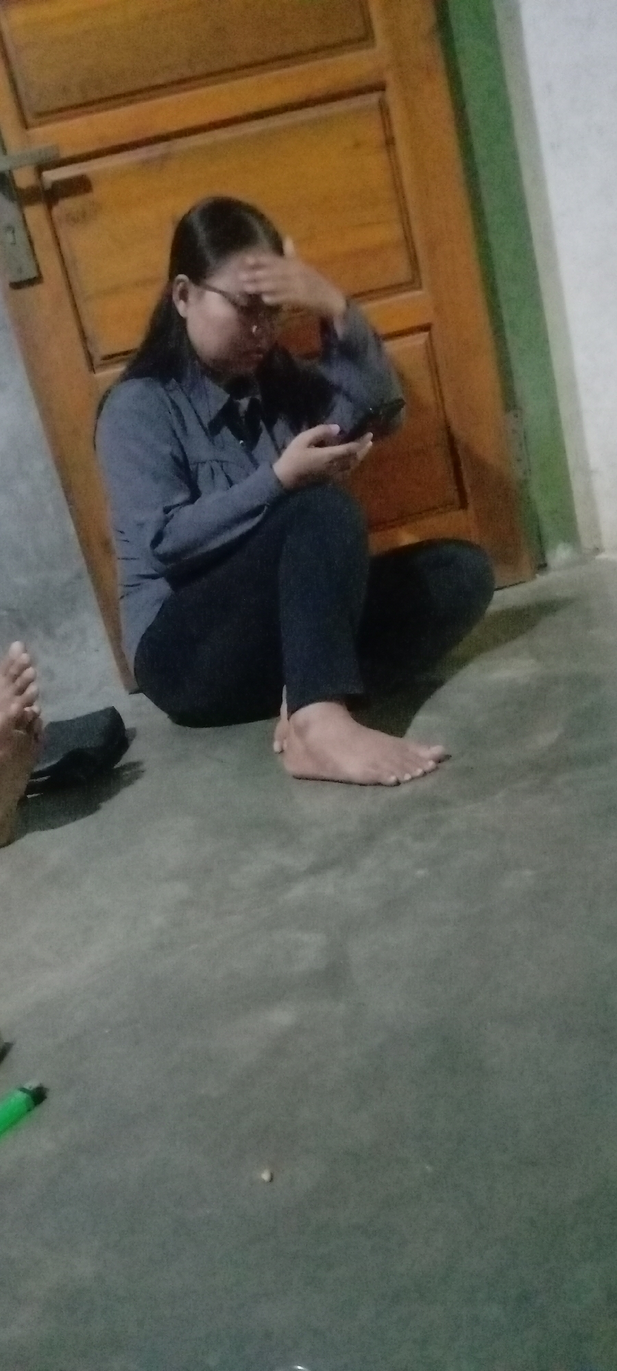 A young woman wearing glasses, a dark blue collared shirt, and dark pants is sitting on the floor, leaning against a wooden door. She is holding a dark smartphone in her right hand and has her left hand on her forehead. Her legs are crossed. To the left of her, another person's bare feet are visible on the floor. The floor appears to be concrete. A dark object, possibly a shoe, is near the feet. A green object, resembling a pen, is on the floor in the lower left corner of the image. The environment suggests an indoor setting, possibly a residential home.