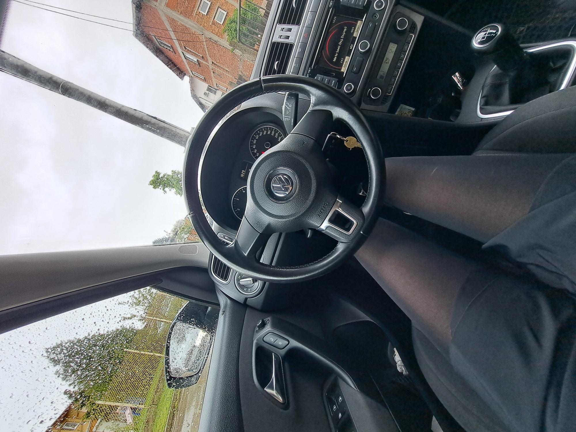 The image provides a tilted interior view from the driver's seat of a Volkswagen car, looking forward through the windshield and to the driver's side. The lower part of a person's legs, clad in dark tights, is visible in the driver's seat.
The steering wheel, featuring a "VW" logo and "AIRBAG" text, is prominent in the foreground. Car keys hang from the ignition near the steering column. Behind the steering wheel, the instrument cluster shows a speedometer marked up to "240 KM/H" with the needle at "0". The infotainment system displays "18:29" (time), the track "Marta Savic-Duj, Duj.mp3", and numbers "2:42" and "-003". Buttons labeled "MEDIA", "RADIO", "PHONE", "SETUP", "SELECT", and "MDI" are also visible. A manual gear stick with a standard 5-speed and reverse pattern is located to the right. The driver's side door panel includes controls for windows and mirrors.
Outside the car, water droplets on the windows indicate rain or recent precipitation, and the sky appears overcast and cloudy. A large utility pole runs diagonally across the view through the windshield. In the background, a multi-story brick building with several windows, along with some trees and fencing, is visible. The car appears to be stationary, as indicated by the speedometer and the keys in the ignition.
