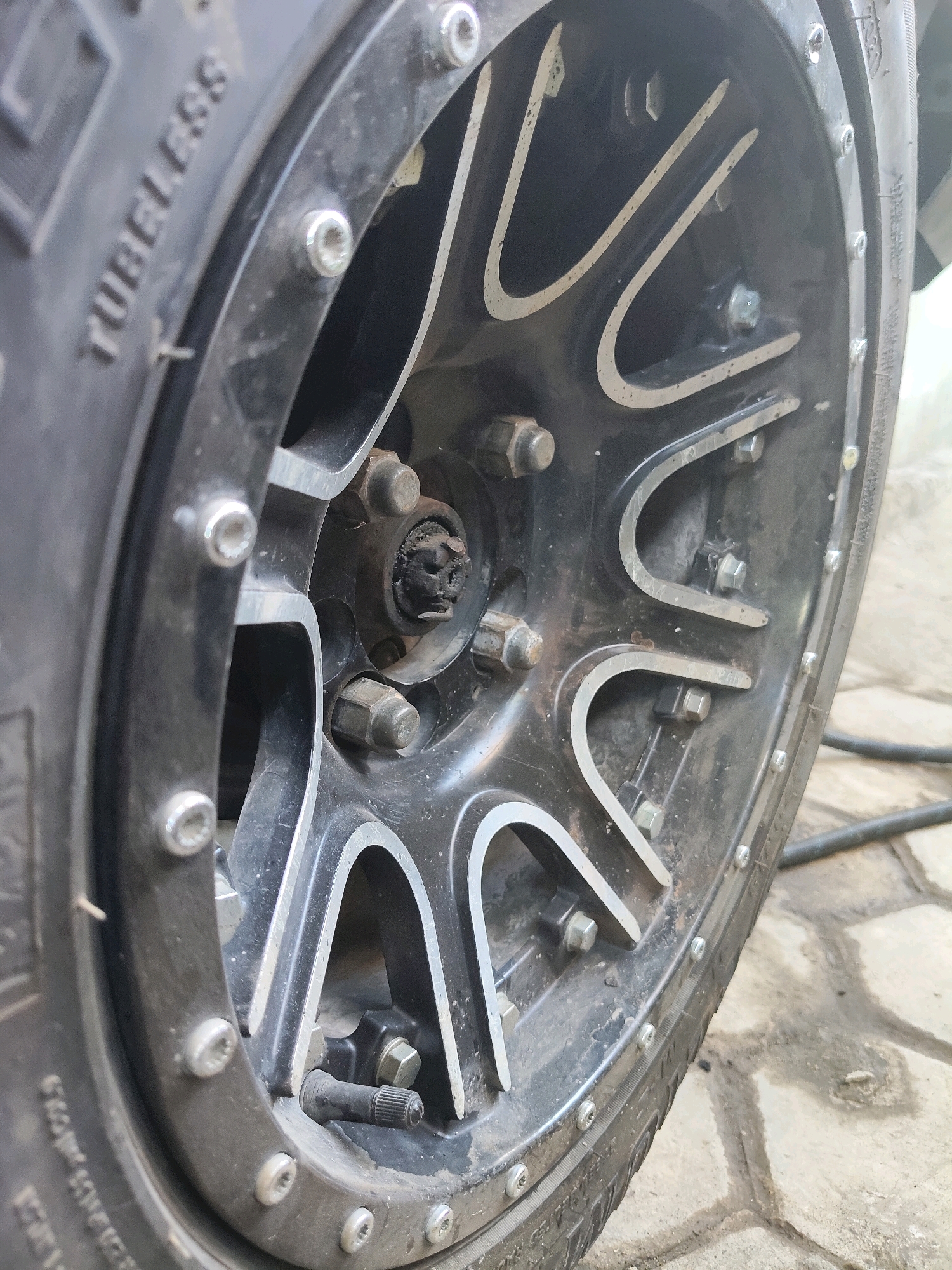 The image shows a close-up, low-angle view of a black and silver alloy wheel of a vehicle, likely a car or truck, parked outdoors on a paved surface with visible grout lines. The wheel is the primary subject, with a black tire featuring the word "TUBELESS" embossed on its sidewall, which is partially visible on the left side of the frame. The alloy rim has a multi-spoke design with silver accents against a black background. Several lug nuts, appearing metallic and hexagonal, are visible securing the wheel to the hub. The central hub area is dark and somewhat obscured by dirt and grime, with a visible nut and what appears to be a cap. A black rubber tire valve stem is also visible protruding from the rim. The lighting suggests it is daytime, with some shadows cast on the ground. The environment is urban, with the paved ground suggesting a street, driveway, or parking area. The presence of a vehicle wheel indicates a setting where vehicles are present. Based on the provided information, this scene is located in Kupang, Indonesia. There are no people visible in the image.