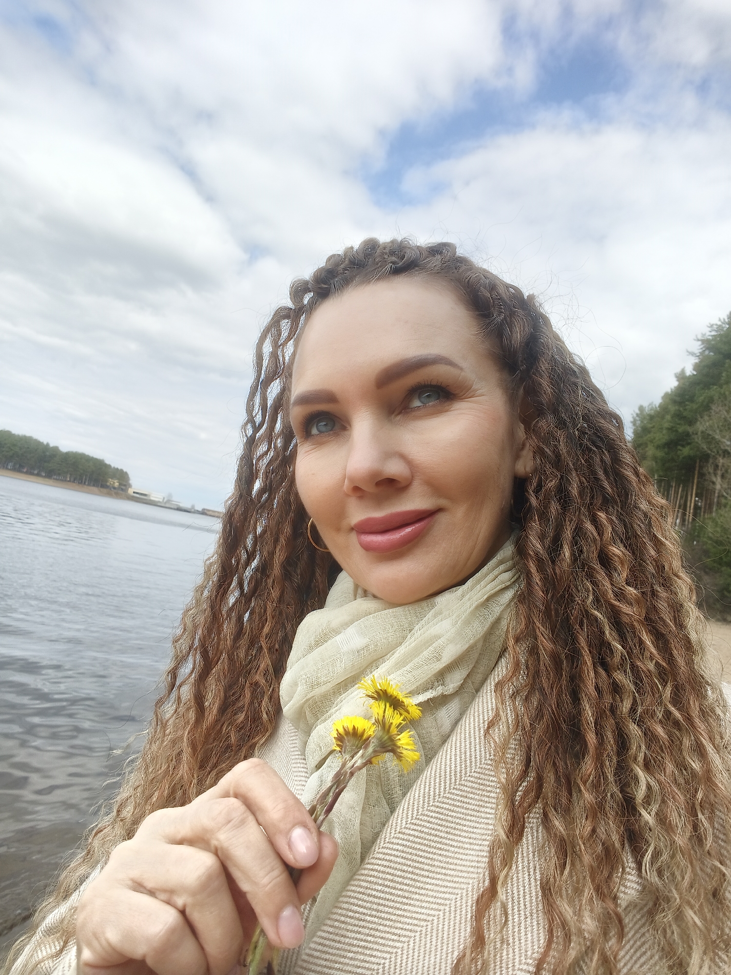 A woman with long, curly, light brown hair stands near a body of water. She is holding three small yellow flowers in her hand. She is wearing a light-colored scarf and a beige patterned coat. In the background, there is a lake, a shoreline with trees, and a cloudy sky. Based on the presence of the Kama River and architectural details visible across the water, the location is likely in or near the city of Chaikovsky, Russia.