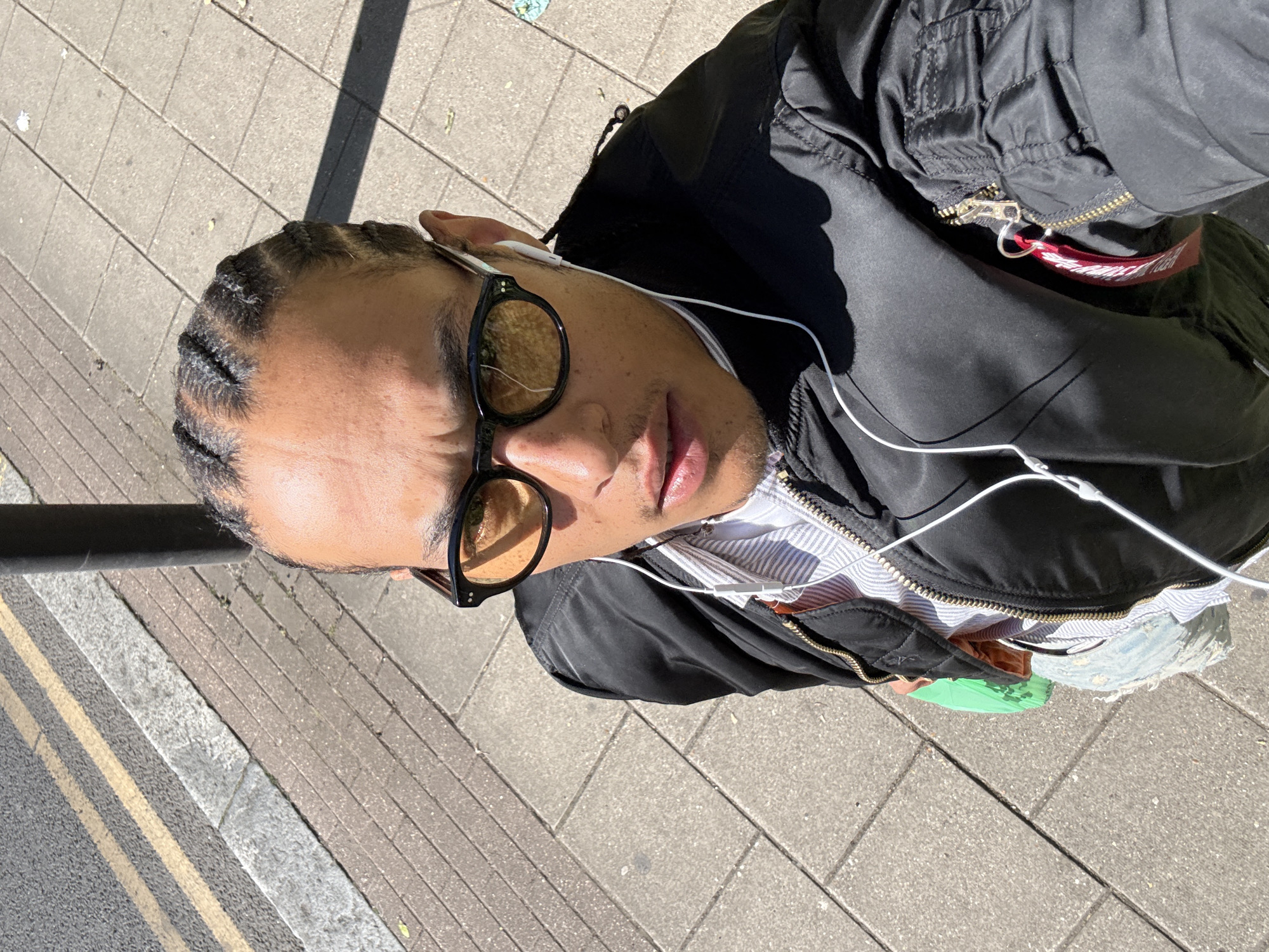 The image presents a high-angle, close-up view of a person outdoors on a sunny day, likely in London, United Kingdom. The subject, a person of color, has tightly braided hair (cornrows) and is looking upwards towards the camera. They wear black-framed sunglasses with yellowish-orange tinted lenses and white wired earphones.
The individual is dressed in a black bomber jacket, unzipped to reveal a white shirt with thin blue or grey stripes underneath. A prominent detail is a red fabric tag, characteristic of flight jackets, attached to a zipper pull on the jacket. The text "REMOVE BEFORE FLIGHT" is clearly visible in white lettering on this tag.
The setting appears to be a sidewalk adjacent to a road. The ground is paved with rectangular grey and brown blocks, while a darker asphalt road section is visible to the left, marked with double yellow lines and a single white line, typical of UK road infrastructure. Strong, defined shadows are cast across the ground and the person's clothing, indicating bright sunlight and a clear sky. A dark, vertical pole is partially visible on the far left edge of the frame. The overall impression is a casual, urban street scene.