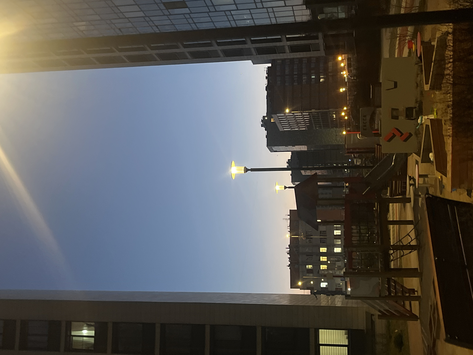 Captured in the city of Saint Petersburg, Russia, the image presents an urban scene at dusk. Flanked by tall buildings with multiple windows, the sky transitions from a deep blue to a lighter gradient. Street lamps illuminate a playground, complete with slides and climbing structures. The playground is located near apartment buildings with illuminated windows, suggesting residential activity. No people are visible in the frame.